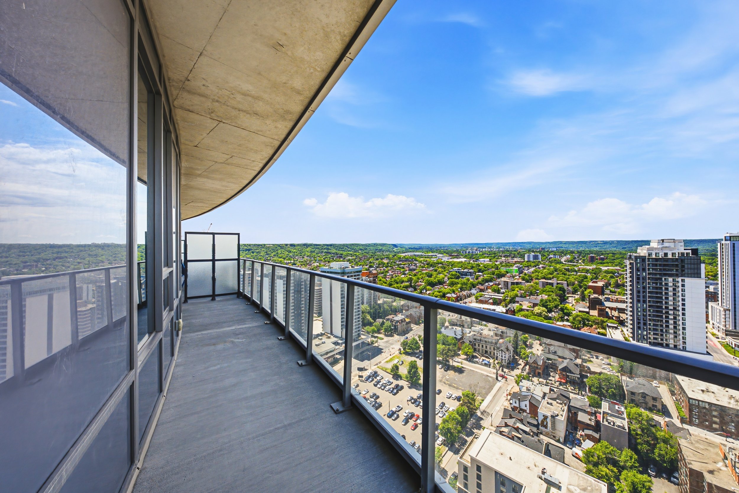 A high-rise balcony with a glass railing overlooks a cityscape with buildings, trees, and streets under a bright blue sky with scattered clouds.