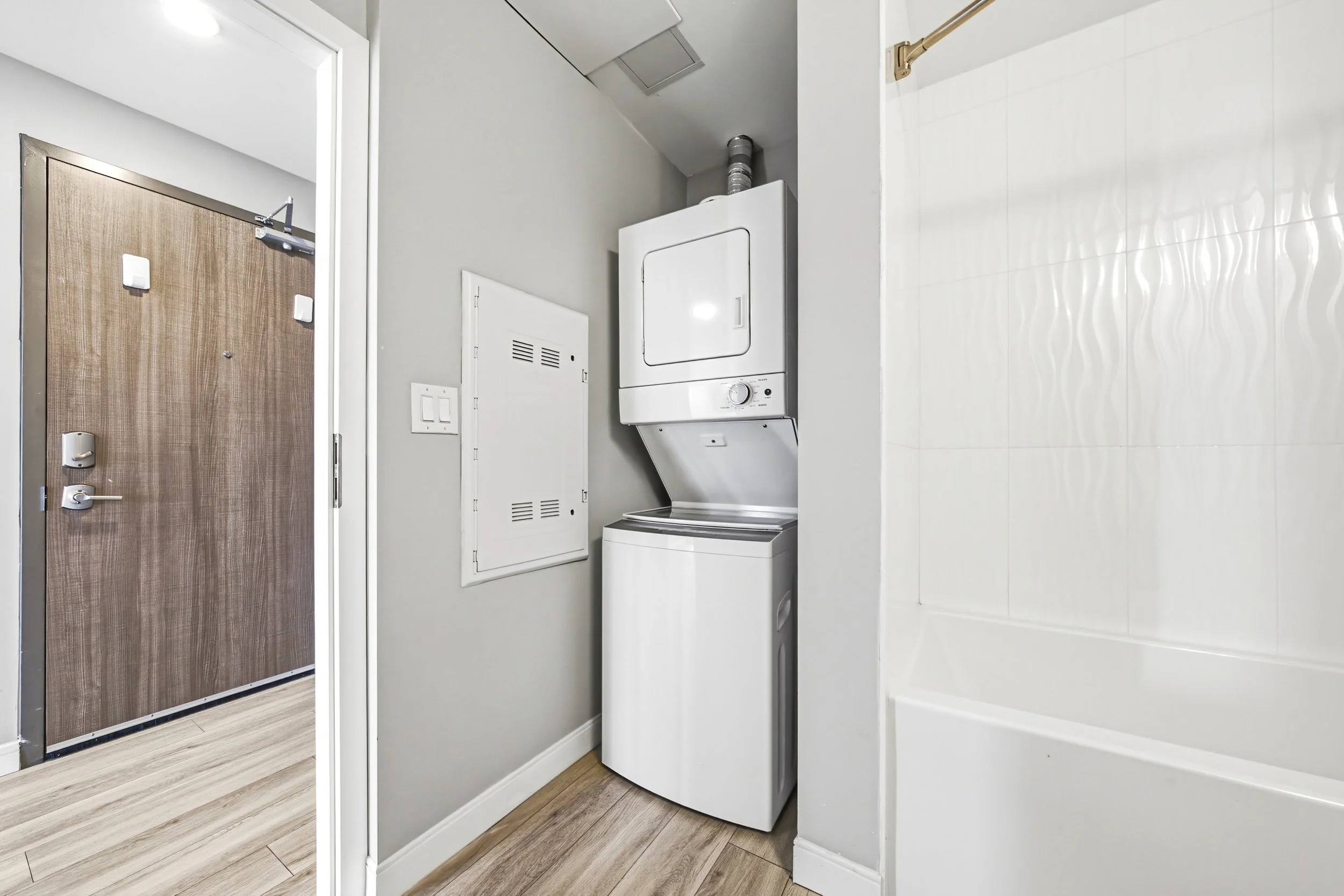 Laundry area with a stacked washer and dryer against a gray wall, adjacent to a white wall with a closed white access panel, and a light wood floor.
