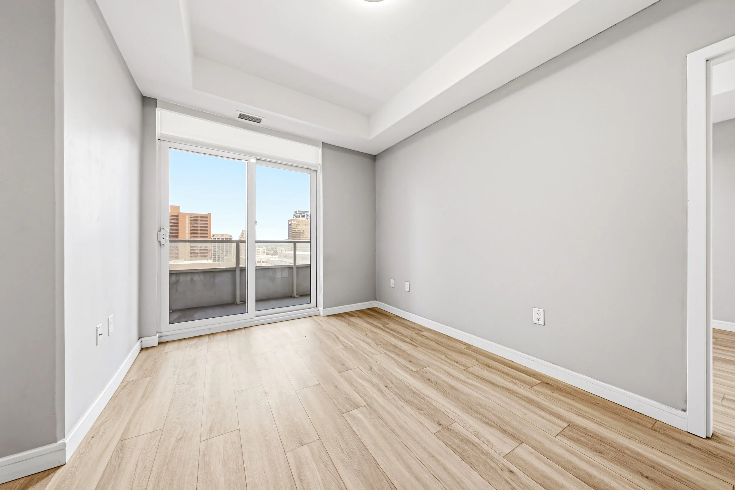 An empty apartment room with wood flooring, white walls, a sliding glass door leading to a balcony, and city buildings outside.