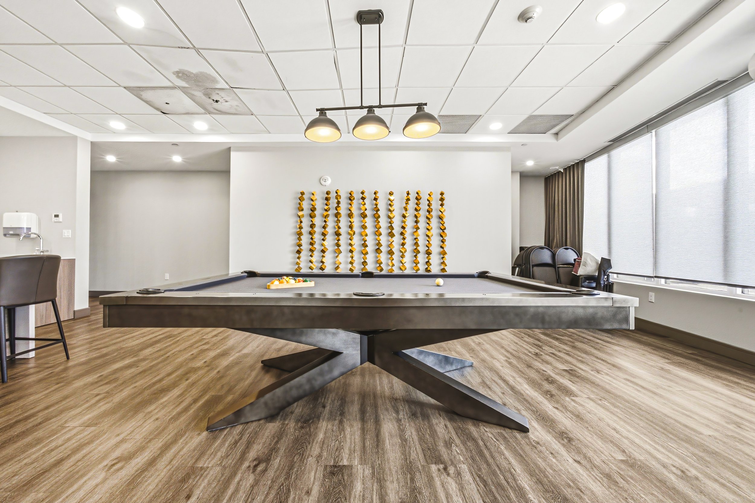 Interior of a recreational room featuring a pool table, wall-mounted yellow decorative elements, chairs near large windows with shades, and a ceiling with recessed lighting and a three-lamp overhead light fixture.