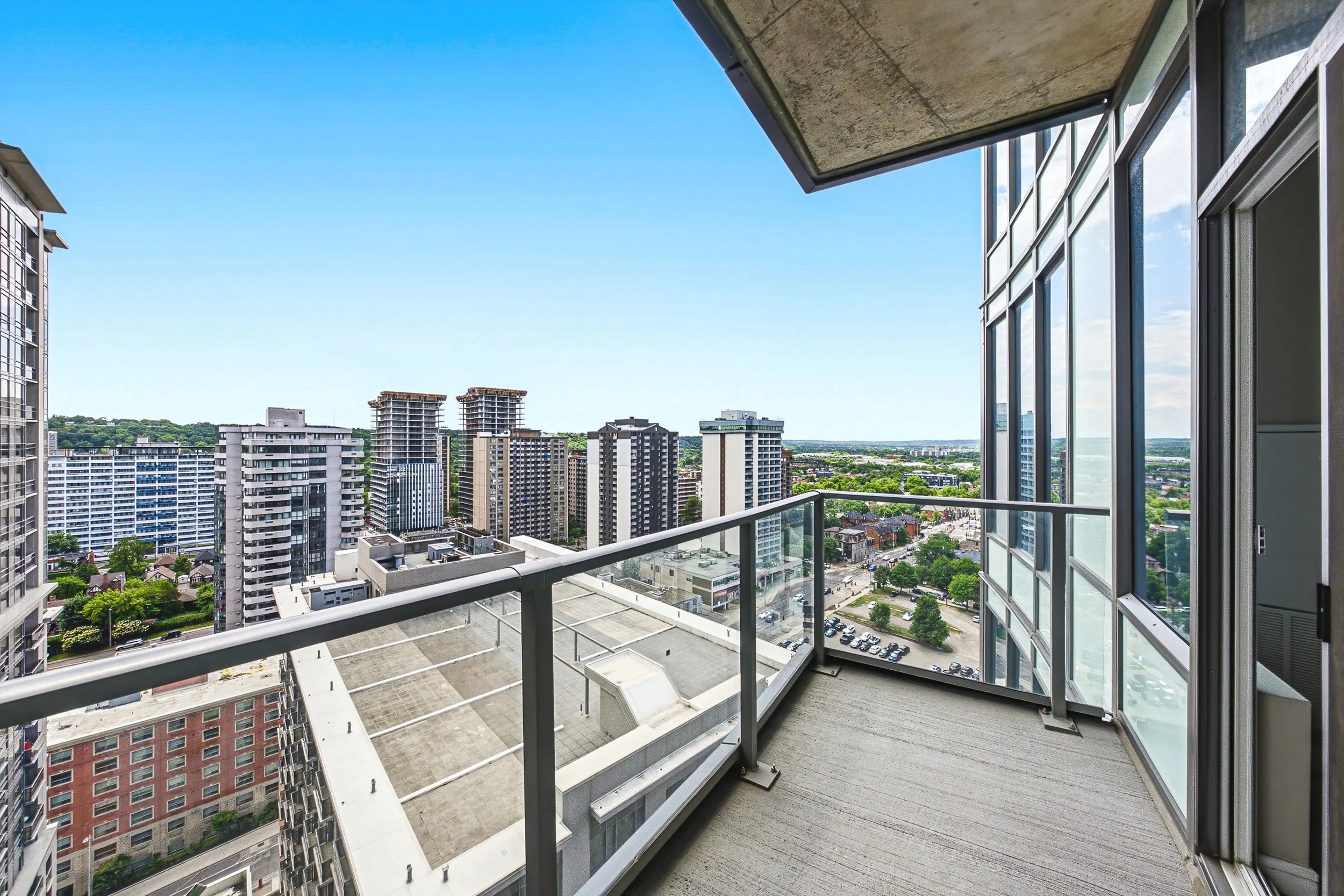 A high-rise apartment balcony with glass barriers, overlooking a cityscape with numerous tall buildings and a clear blue sky.