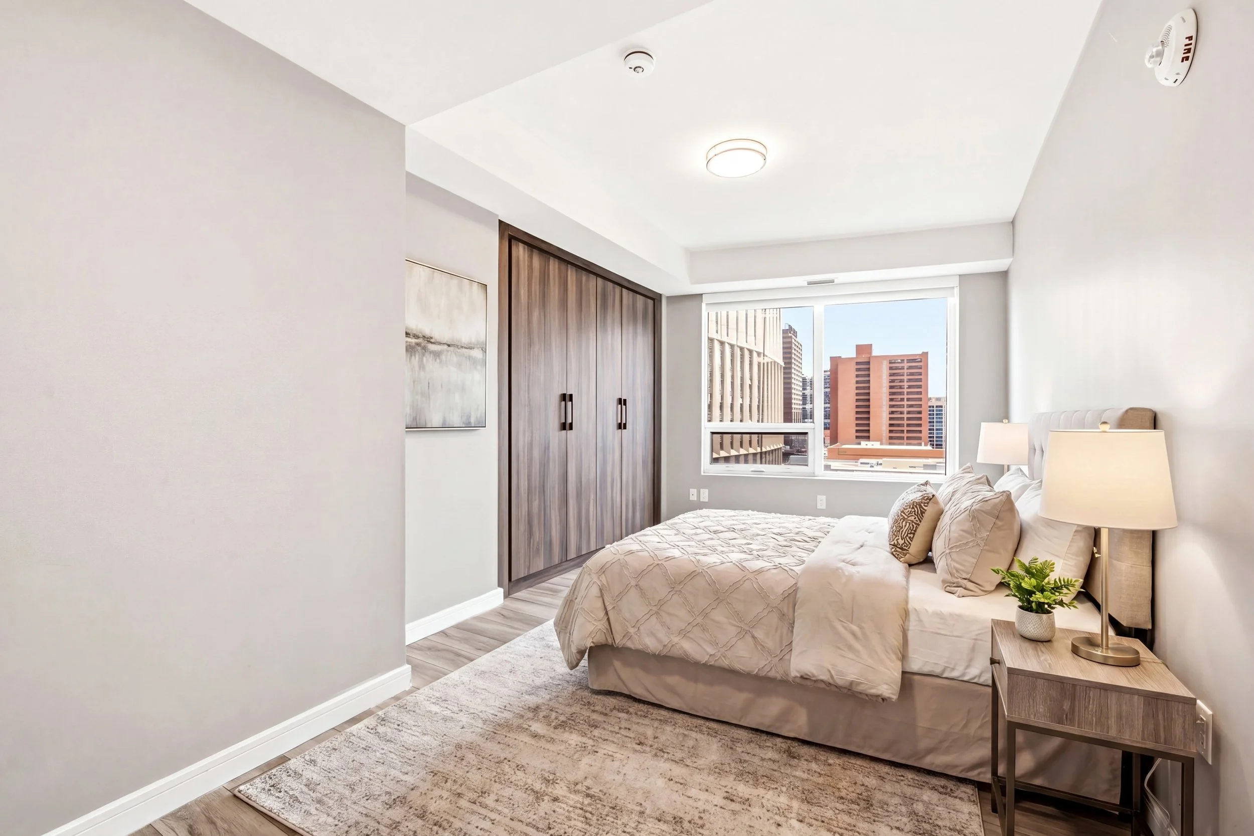 A modern bedroom with a large window overlooking city buildings, a beige bed with pillows, a wooden bedside table with a lamp and a plant, and a wooden closet.