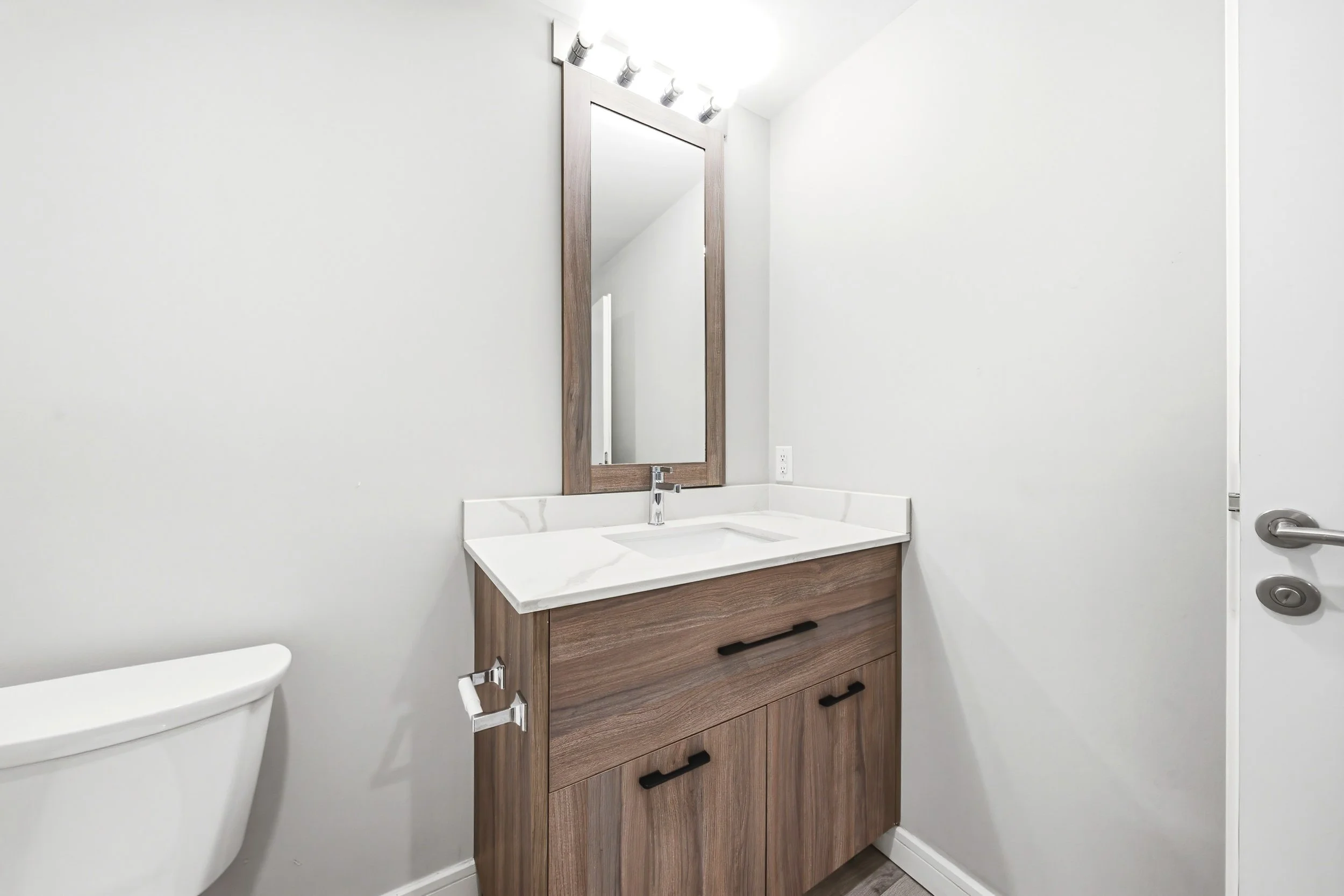 A modern bathroom with a wooden vanity with black handles, a rectangular mirror, and a white countertop with a built-in sink. Part of a toilet is visible on the left, and there are three circular fixtures on the electric hand dryer on the right.