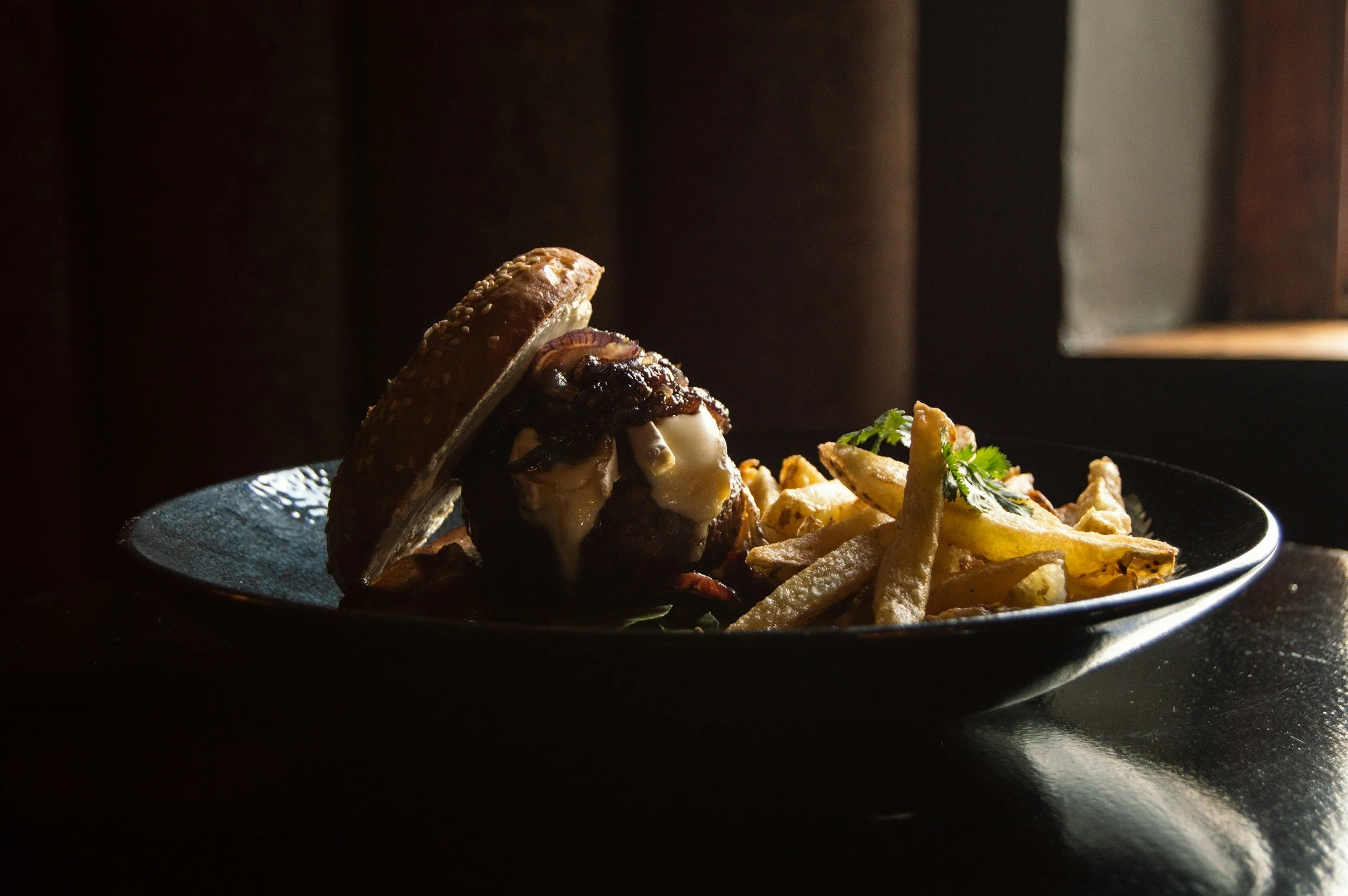 A plate with a burger topped with cheese, mushrooms, onions, and sauce, served with French fries, garnished with a sprig of cilantro, set against a dark, low-light background.