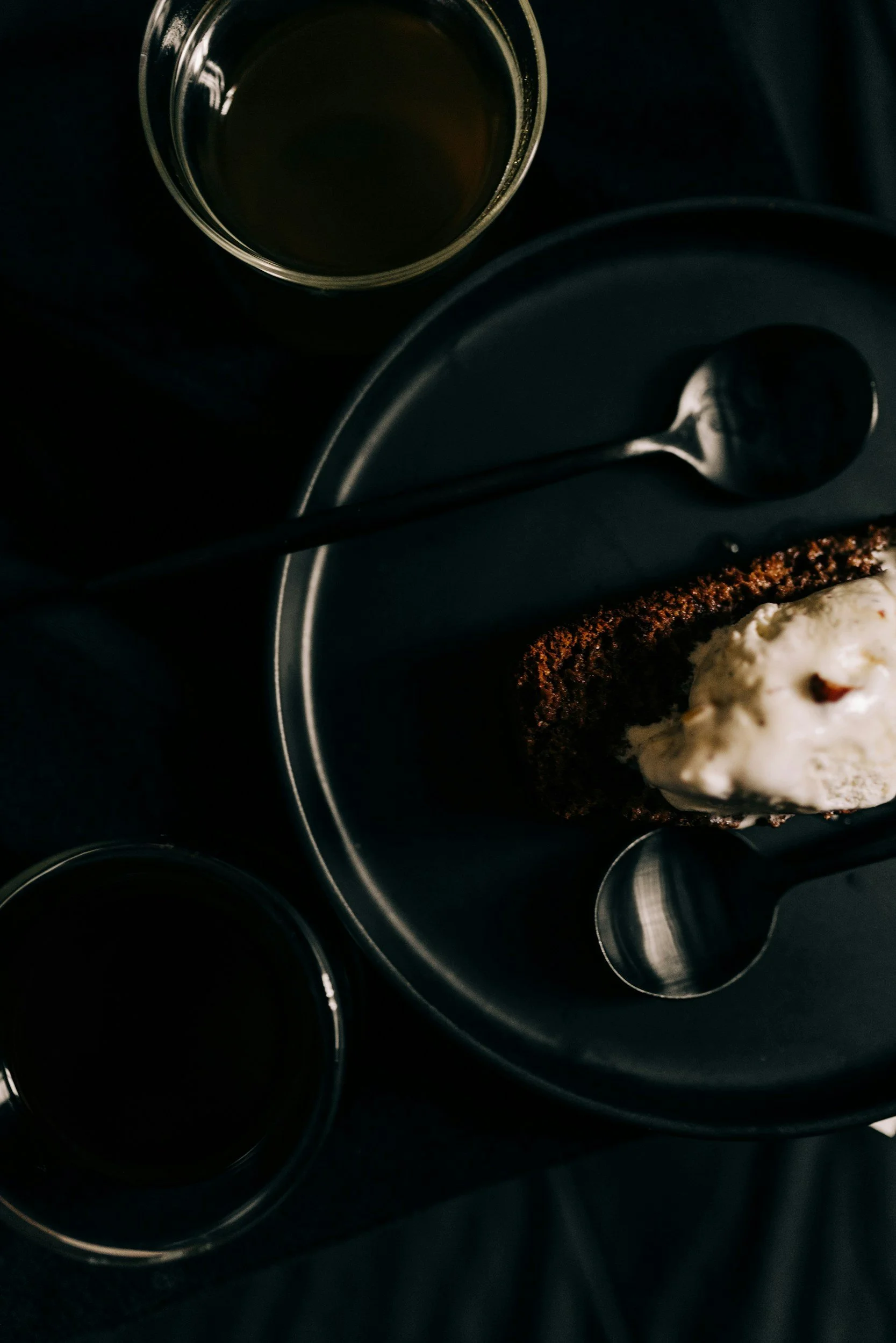 A top view of a dark-colored plate with a slice of chocolate cake topped with whipped cream, a spoon, and a cup of coffee on a black surface.