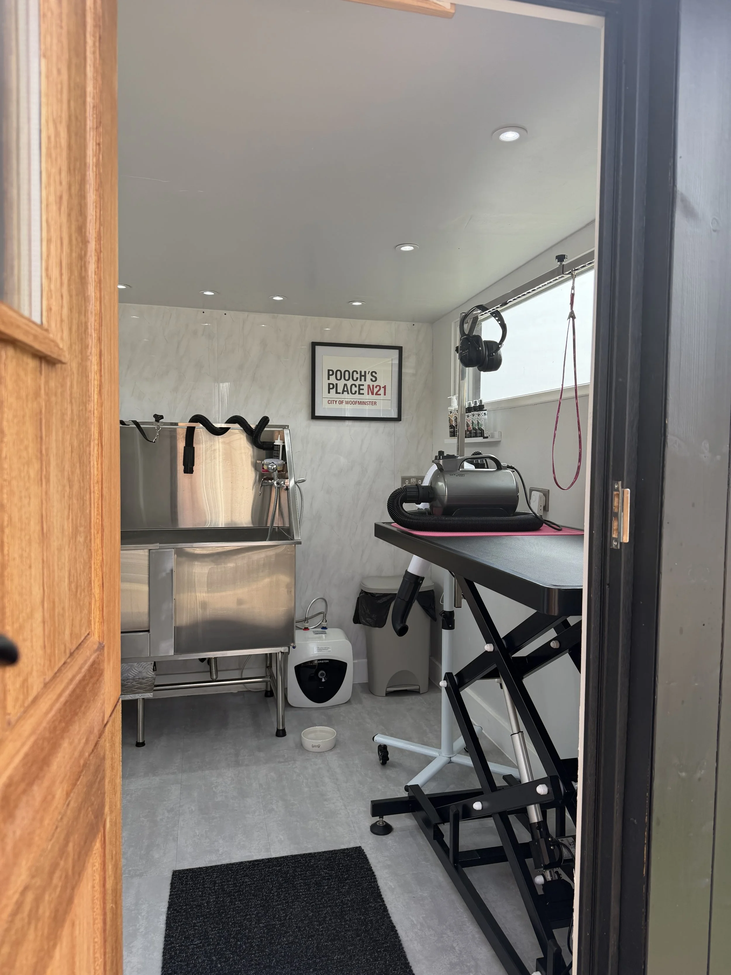 View of a pet grooming station through a doorway, featuring grooming equipment, a framed sign reading 'Pooch's Place N21 City of Woofminster,' a grooming table, and various tools. The room has light-colored walls, a window with blinds, and ceiling lights.