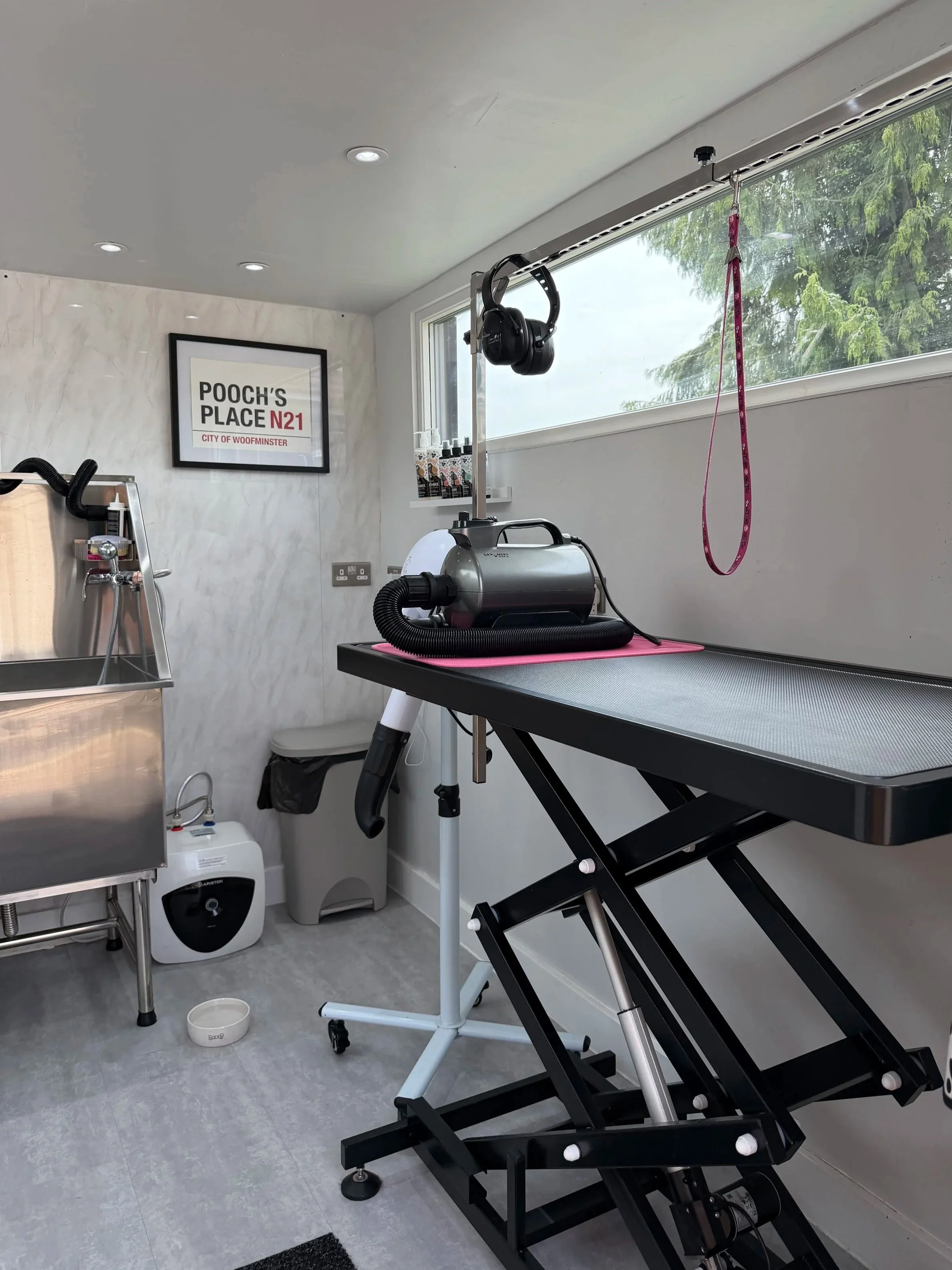 A grooming or veterinary room with an adjustable grooming table, a black head gadget hanging from the ceiling, a pink leash, a grooming spray, a stainless steel sink, a white towel, a small waste bin, and wall art reading 'Pooch's Place N21, City of Woominster'.