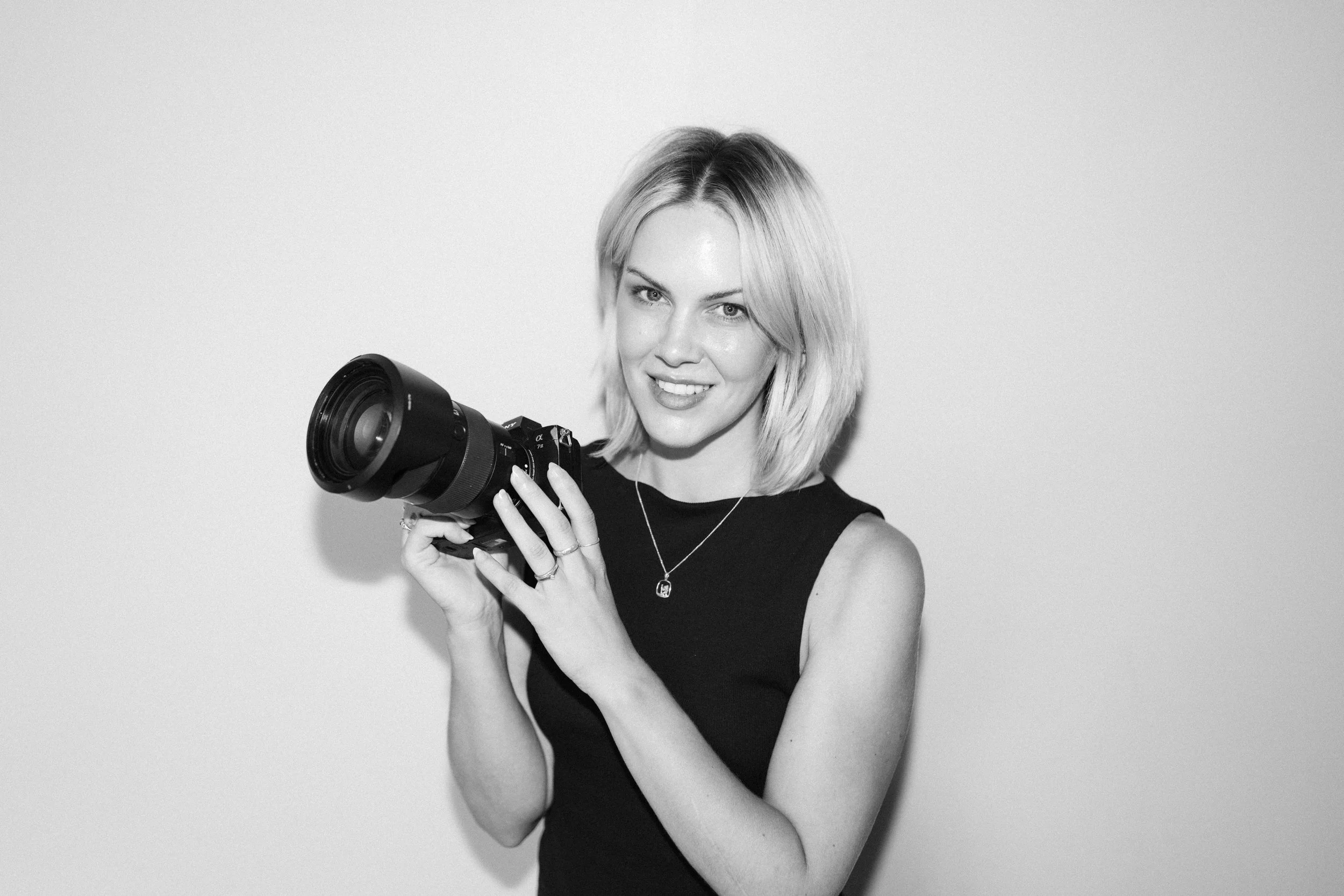 Black and white photo of a woman with shoulder-length blonde hair, holding a camera in front of her face, looking at the camera with a slight smile, wearing a sleeveless black top and a necklace.