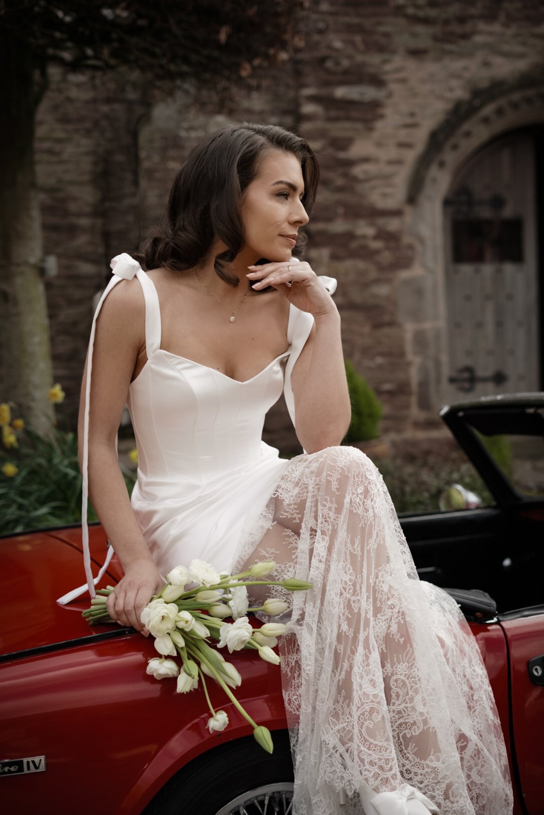 A woman in a white wedding dress sitting on a red convertible car holding a bouquet of white flowers with a lace detail. She is sitting outdoors next to a stone building and a tree.