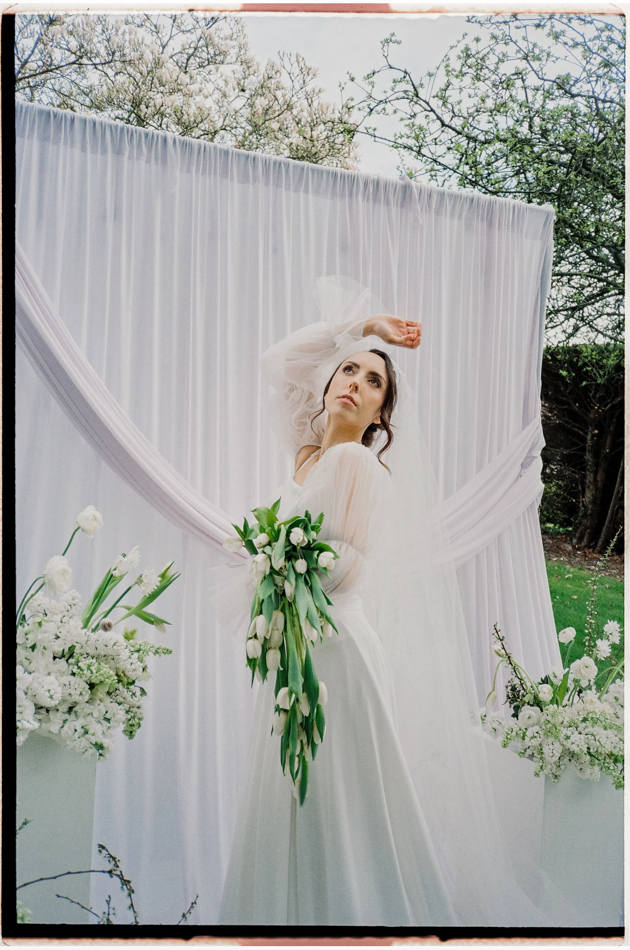 A woman dressed in a white wedding gown holding a bouquet of white tulips and roses, standing in front of a white draped backdrop outdoors during spring, surrounded by white floral arrangements and greenery.