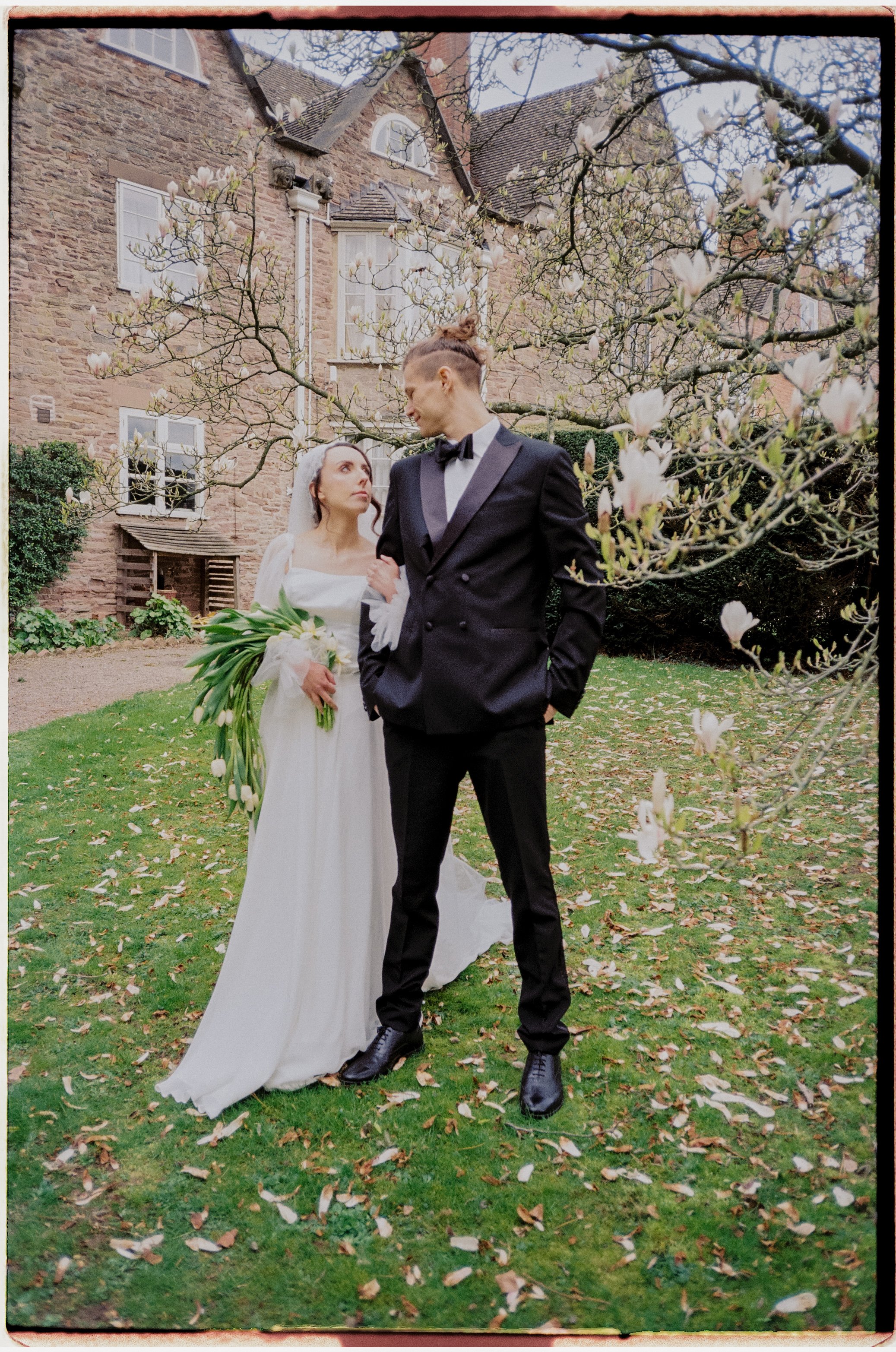 A bride and groom standing outdoors near a blooming tree with a brick house in the background. The bride is holding a bouquet of white flowers and wearing a white dress, while the groom is dressed in a black tuxedo with a bow tie, looking at the brid