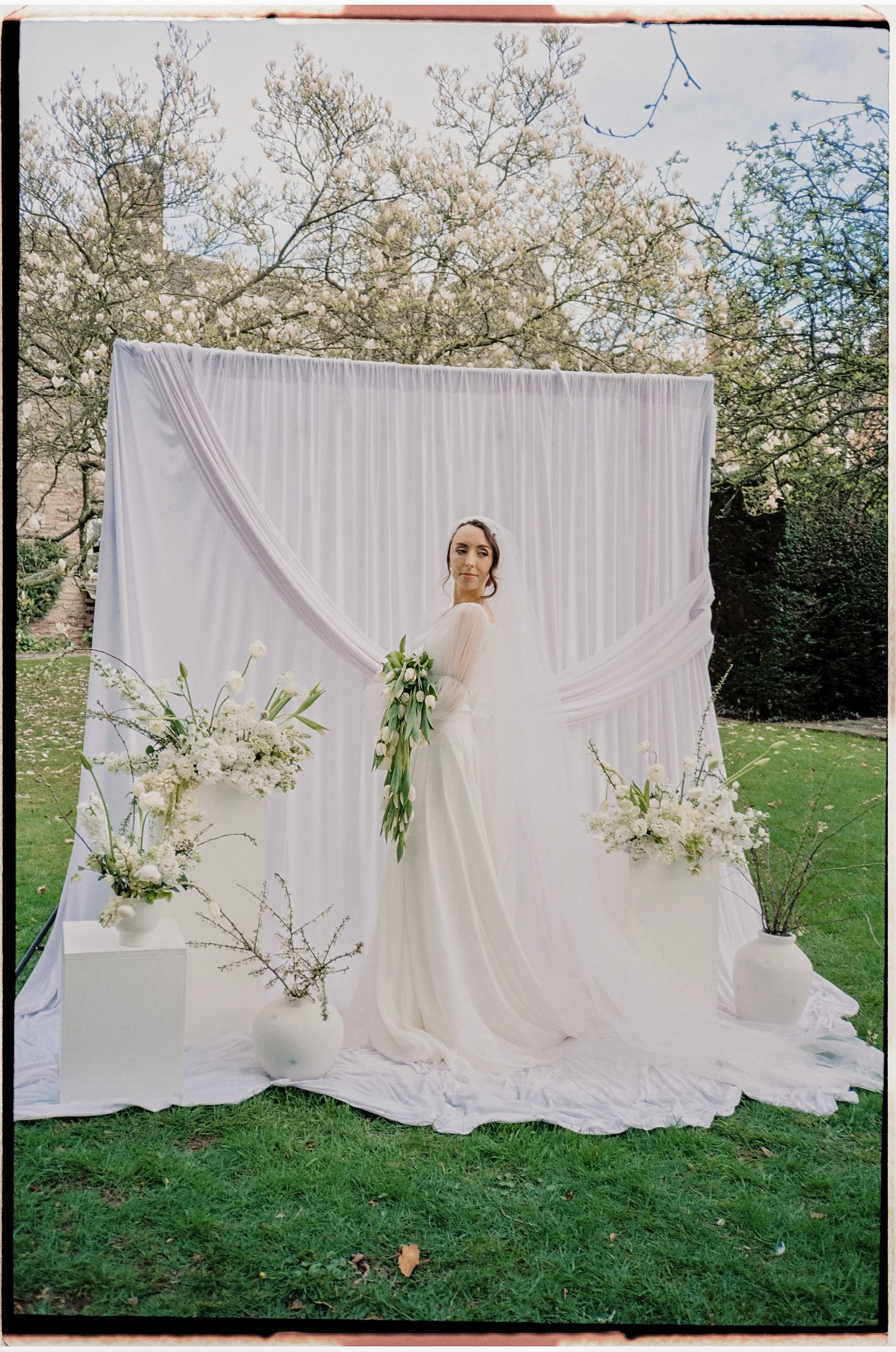 A woman dressed in a white gown holding a bouquet of white lilies, standing in front of a white draped backdrop with floral arrangements on white pedestals outdoors, surrounded by trees with budding leaves.