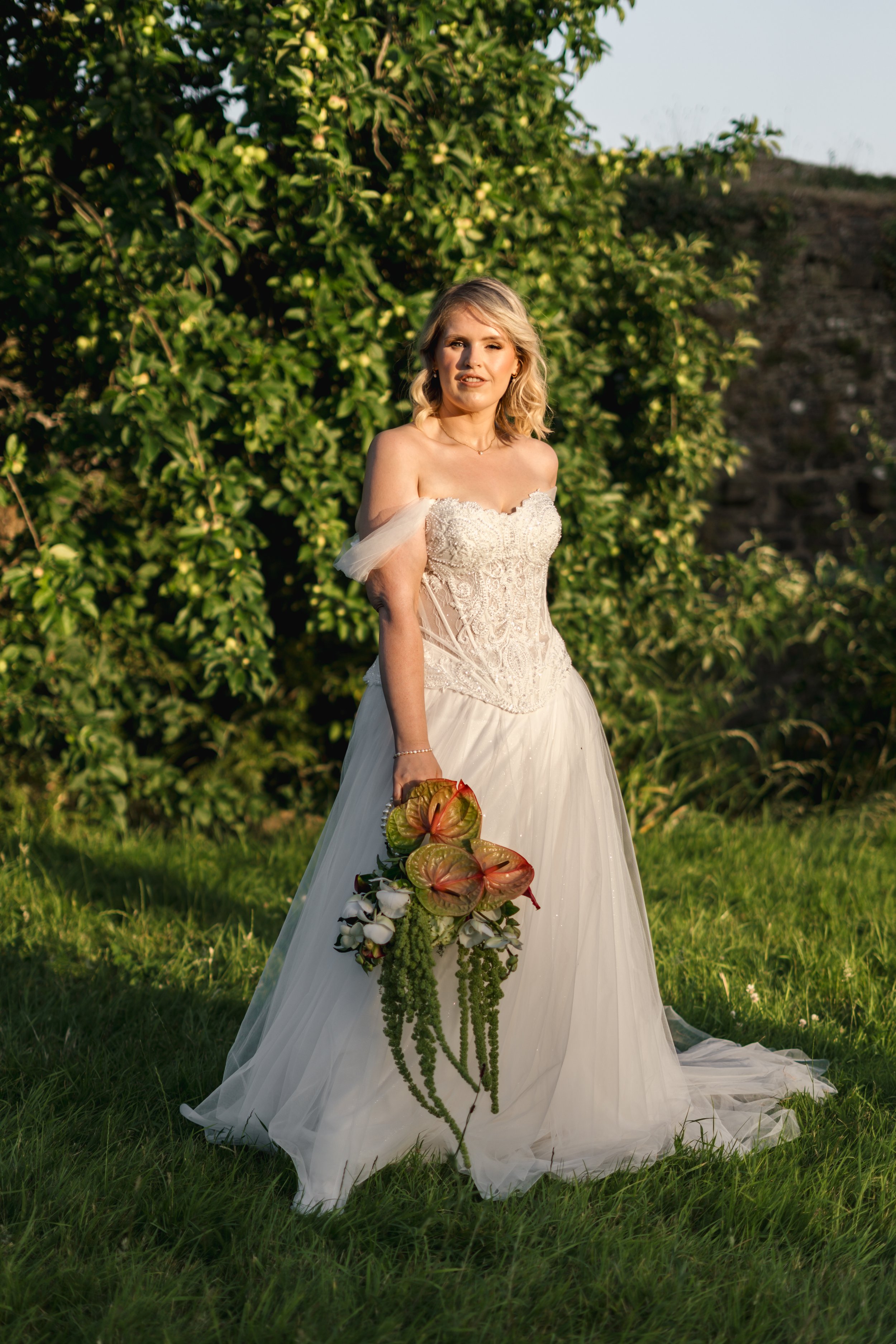 A woman in a wedding dress holding a bouquet of flowers stands outdoors on grass with green trees in the background.