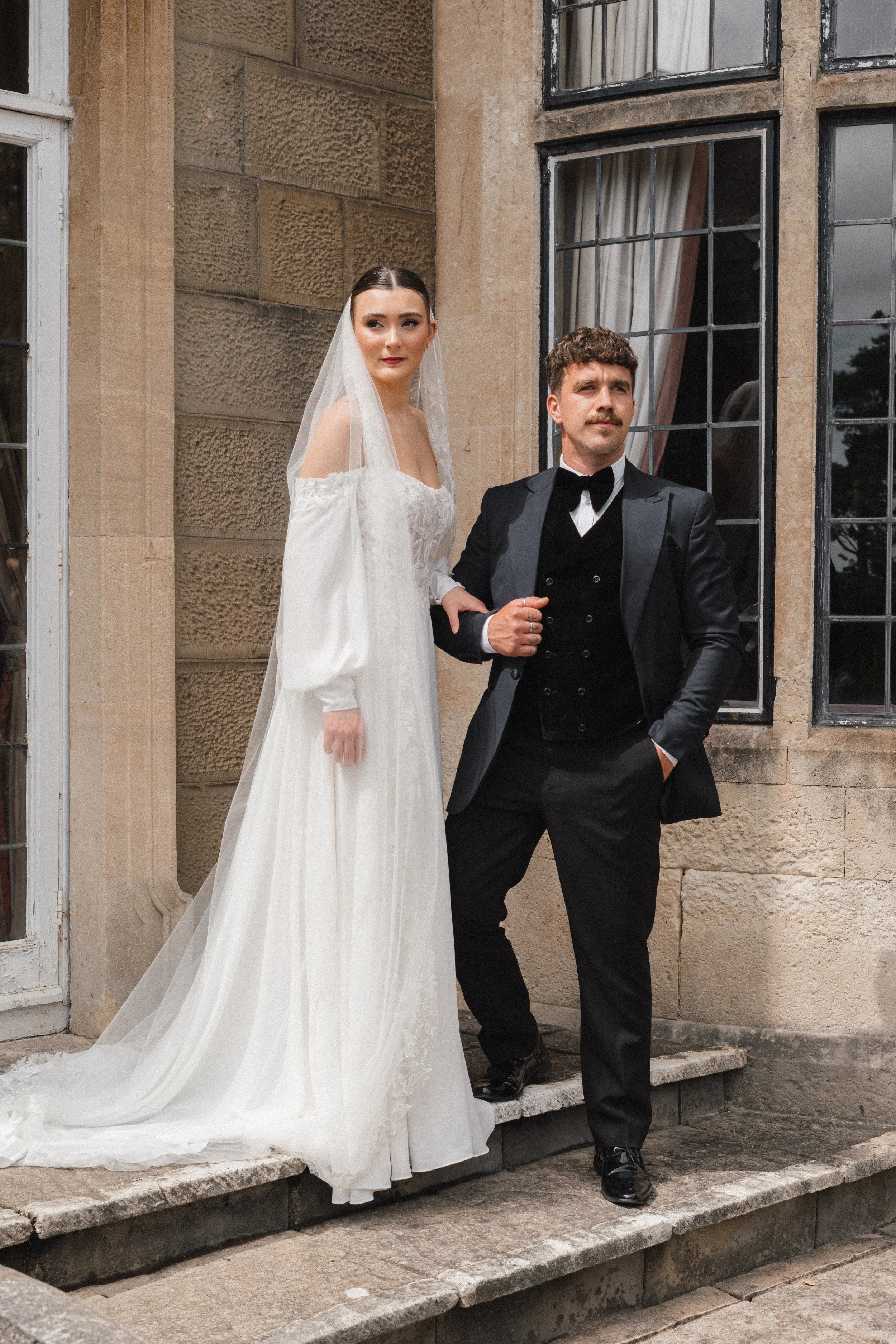 A bride in a white gown with long flowing skirt and veil standing on steps outside a stone building, arm-in-arm with a groom in a black tuxedo with bow tie and mustache.