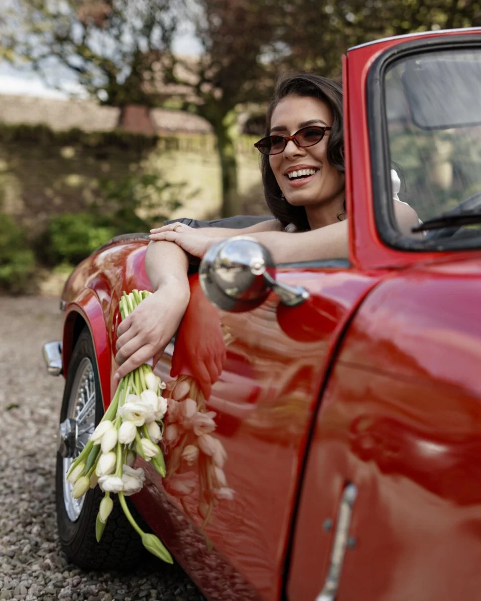 A woman with dark hair wearing sunglasses and a white top, smiling and leaning on the door of a red vintage convertible car, holding a bouquet of white flowers. The background includes trees and a stone wall.