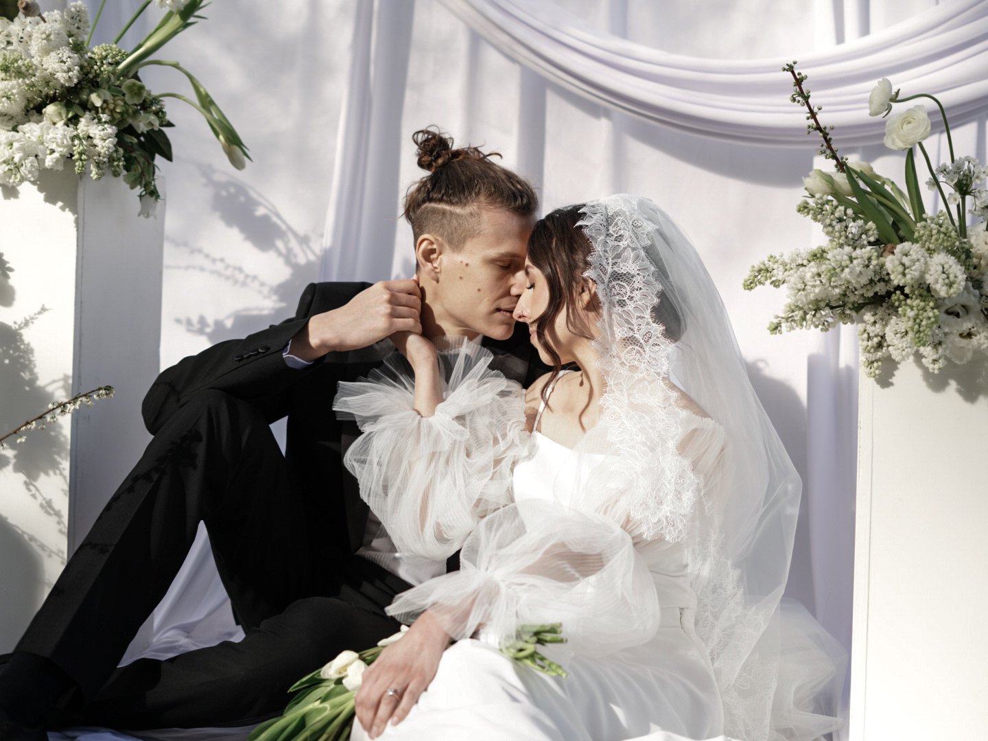 A bride and groom are sitting very close together, touching foreheads, holding each other's hands, with a white floral backdrop and draped fabric.