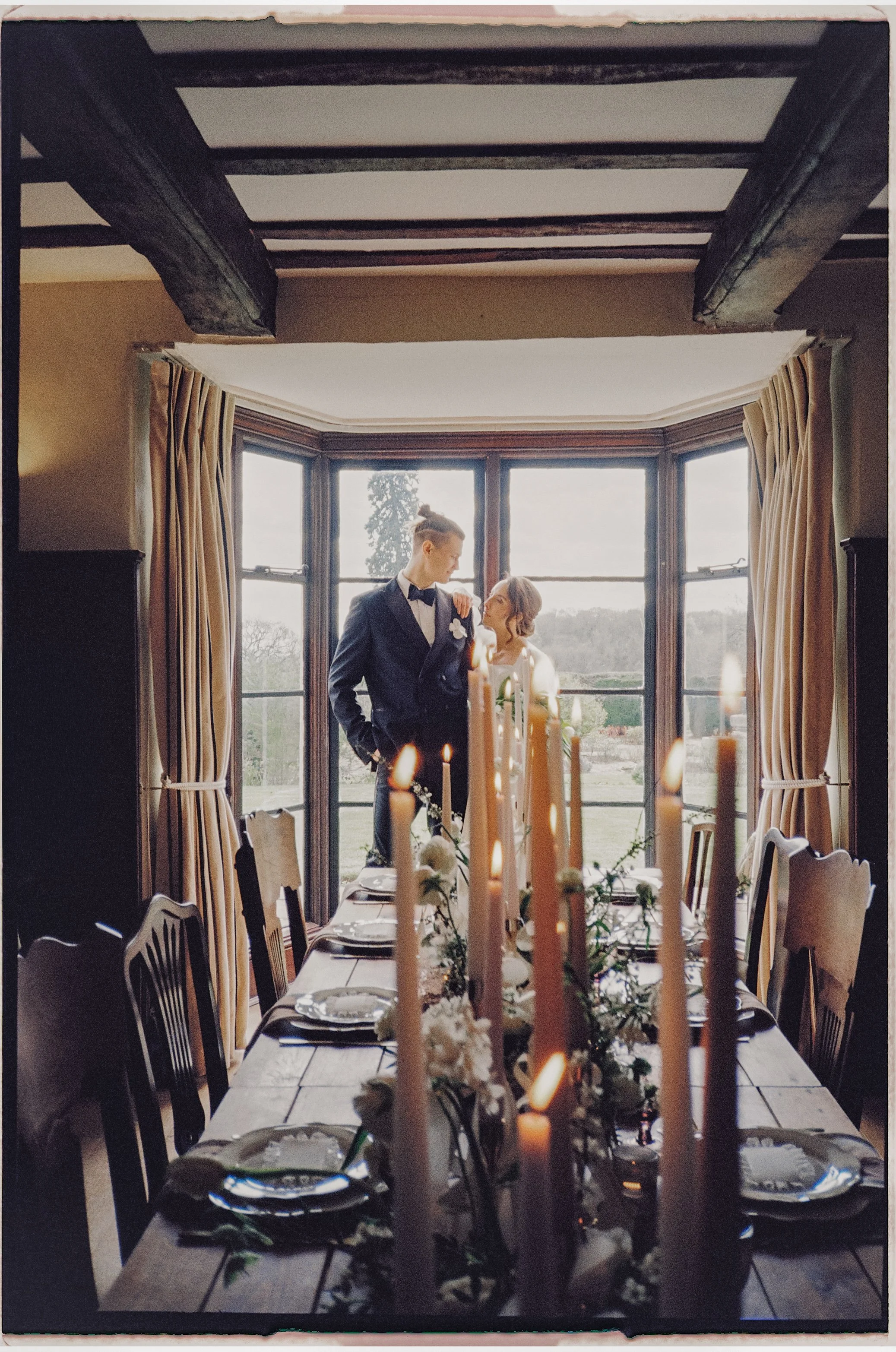 A dining room with a long table set with plates, glasses, and floral centerpieces, illuminated by candlelight. There is a large bay window with curtains, and a wedding couple stands outside the window, with the groom in a tuxedo and the bride in a wedding dress, sharing an intimate moment.