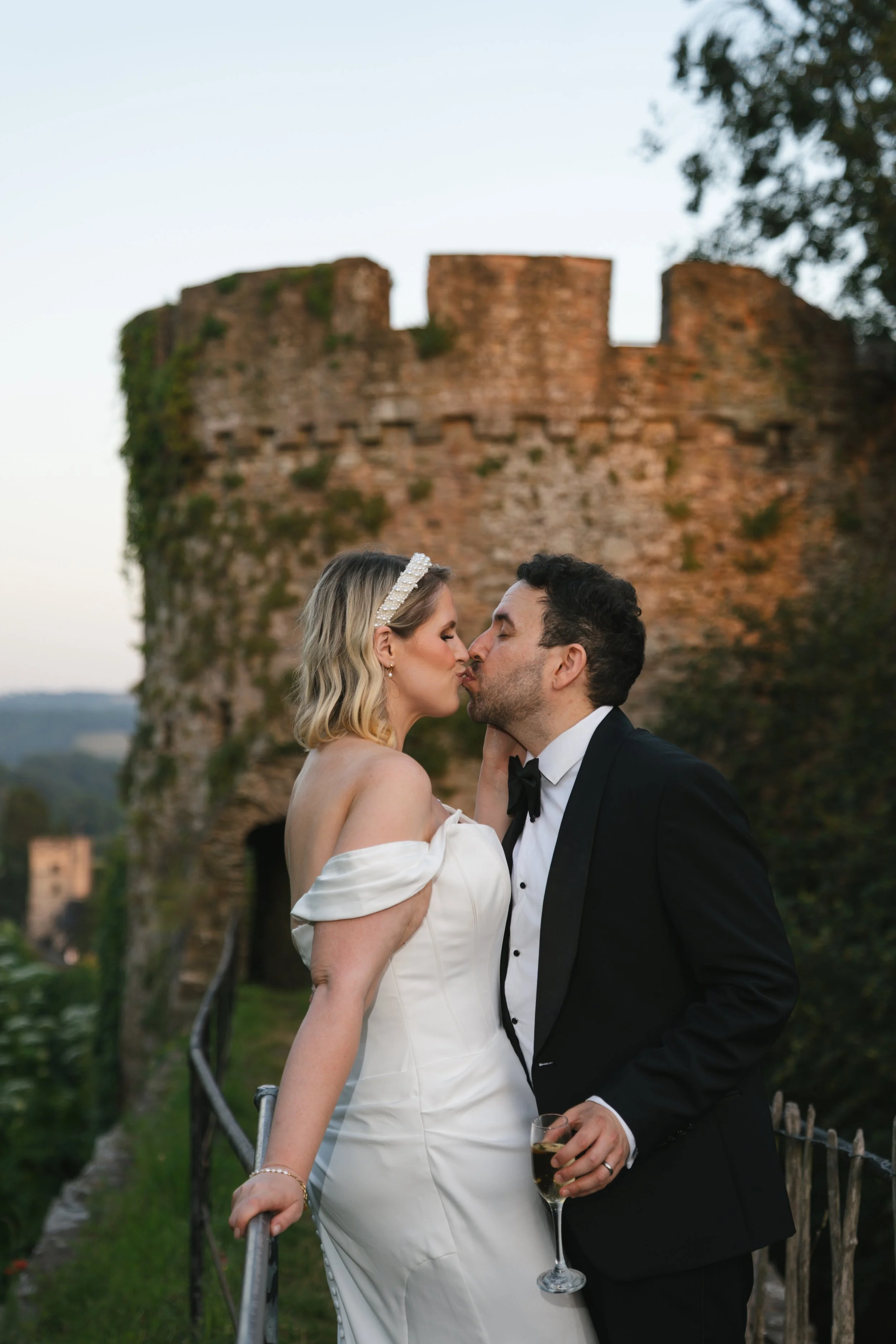 A bride and groom kissing outdoors, with an ancient stone castle in the background, the bride wearing a white off-the-shoulder wedding dress and a pearl headband, holding a glass of champagne, and the groom in a black tuxedo holding a champagne glass