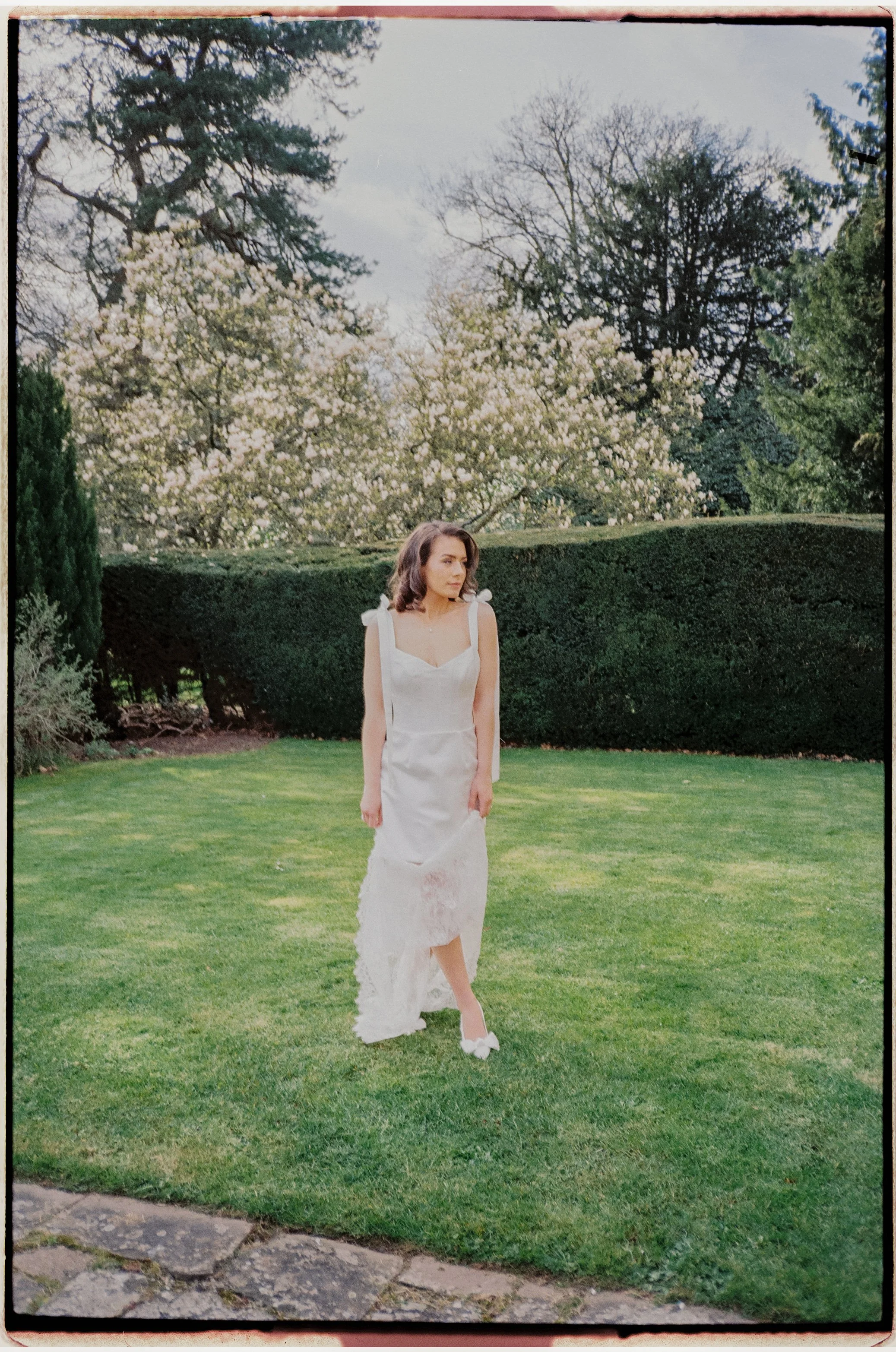 A woman in a white dress walking on grass in a garden with trees and bushes in the background.