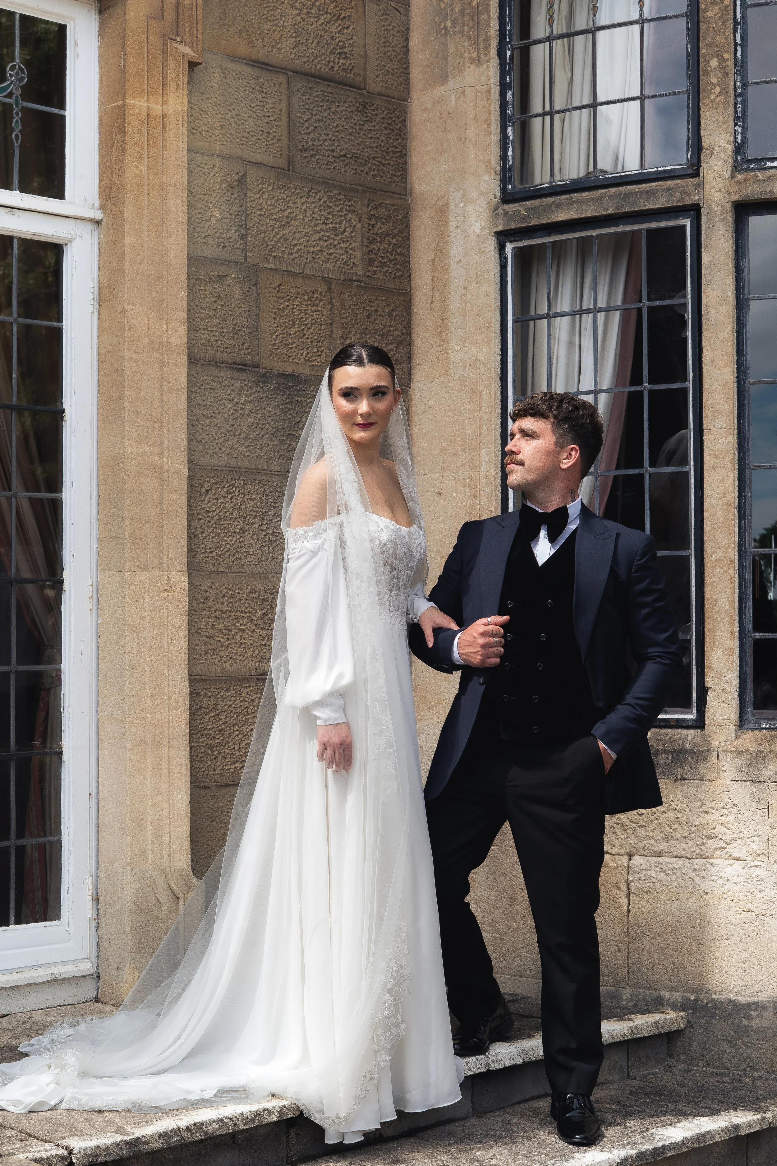 A bride in a white wedding gown with a long train and a veil, standing outside a building, holding hands with a groom in a black tuxedo, near a window with black frames and beige stone walls.