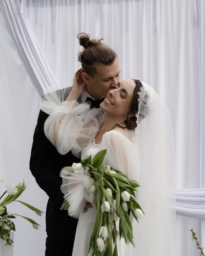 A happy couple on their wedding day, with the groom kissing the bride on the head, holding a bouquet of white tulips.