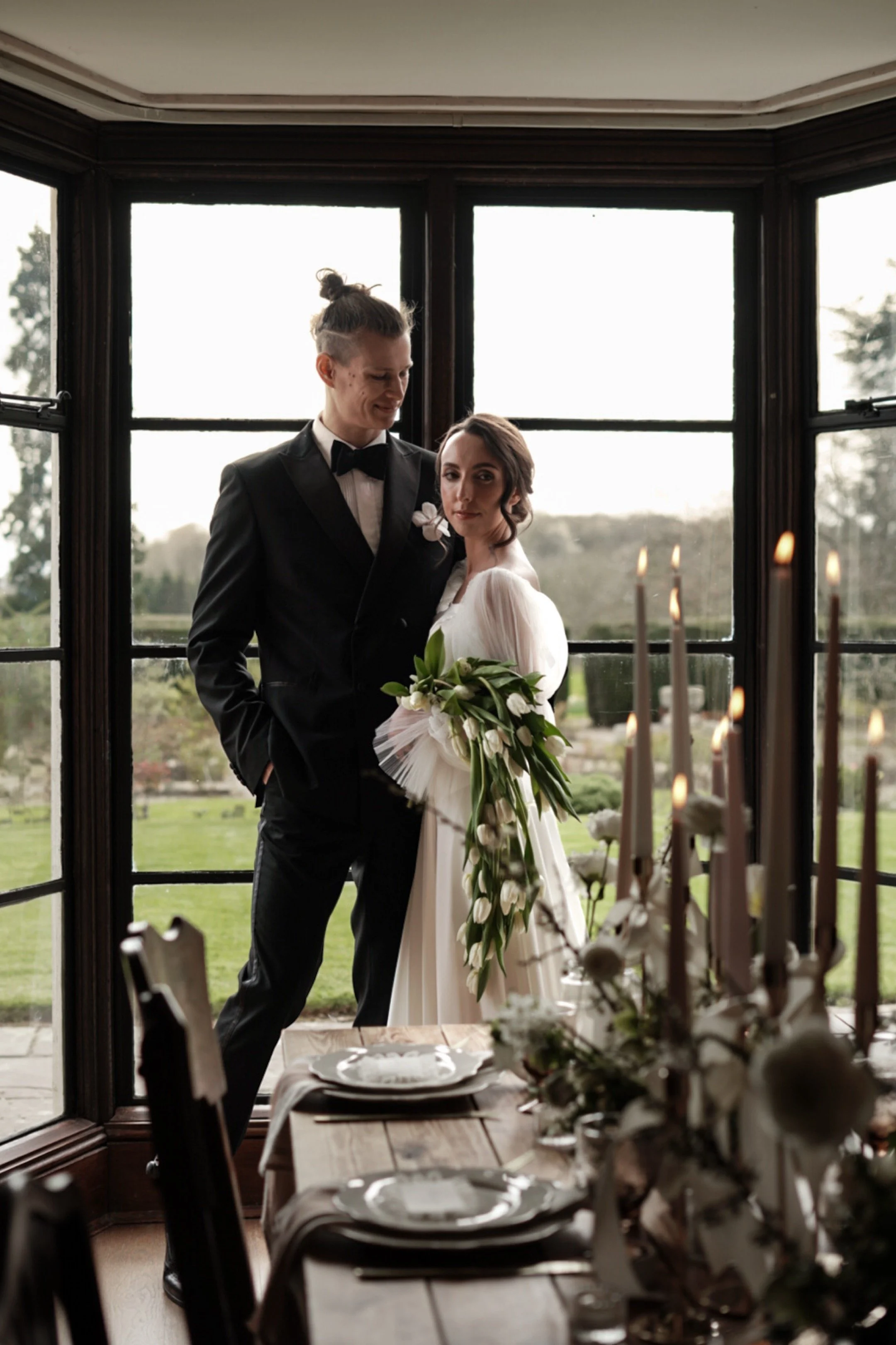 A bride and groom standing inside a room with large windows, outdoors scenery in the background, dining table with candles and floral centerpiece in the foreground.