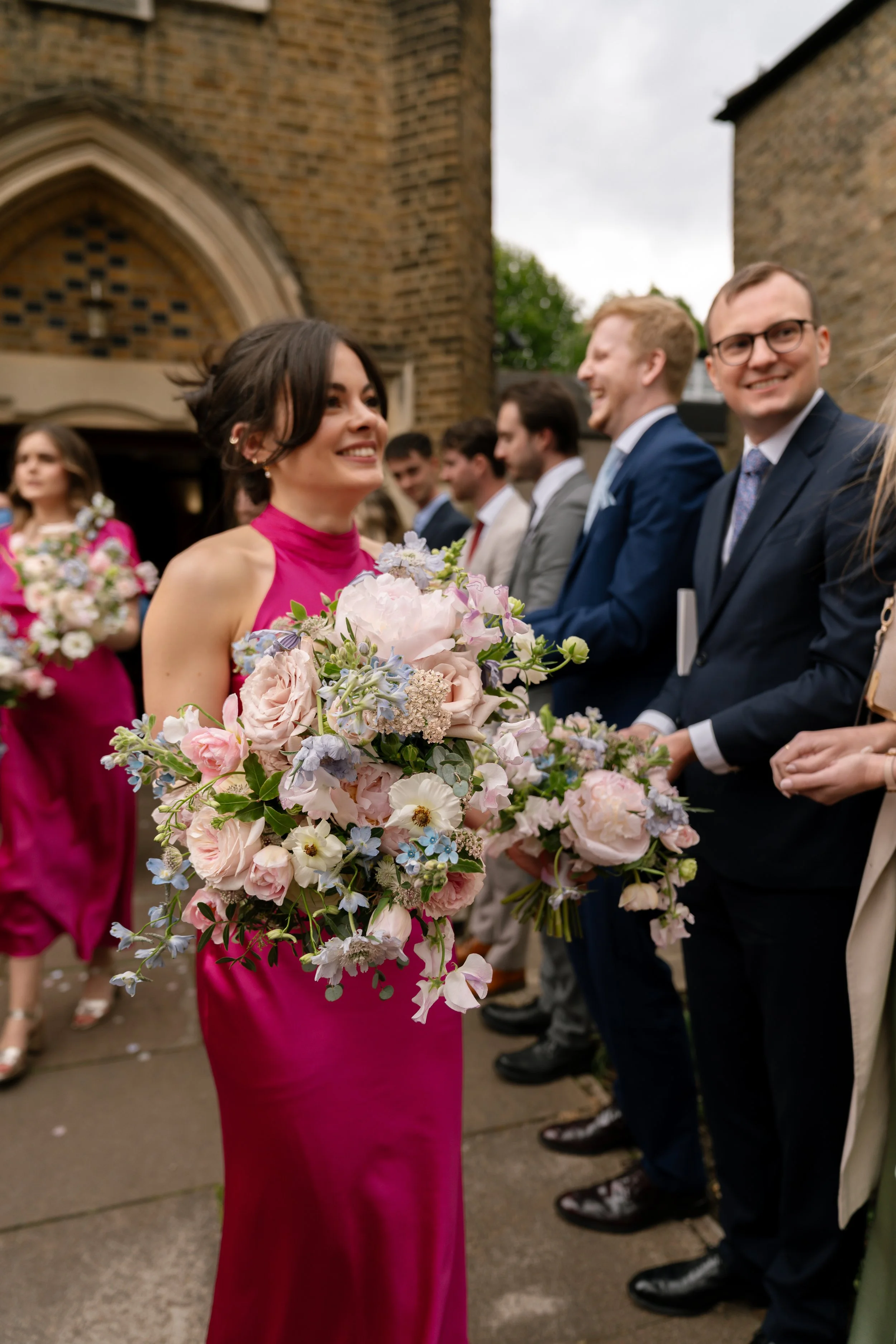 A woman in a pink dress holding a bouquet of flowers at a wedding or celebration, with people in suits standing behind her in front of a brick building.