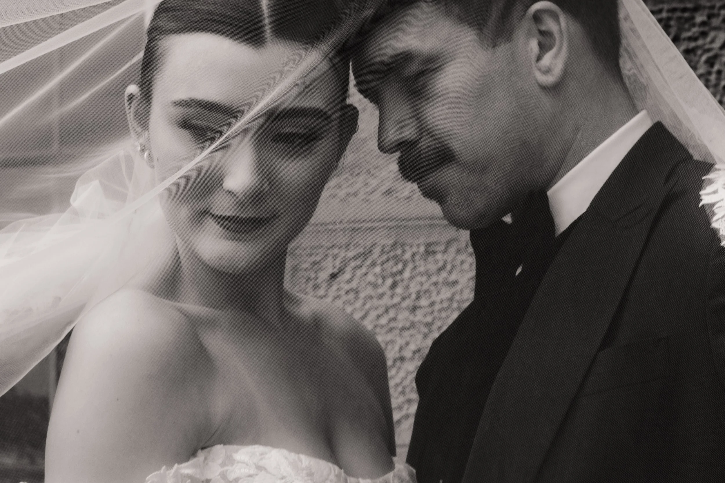 Black and white photo of a bride and groom close together, with the bride wearing a veil, earrings, and a strapless dress, and the groom in a suit and bow tie, both with serious expressions.