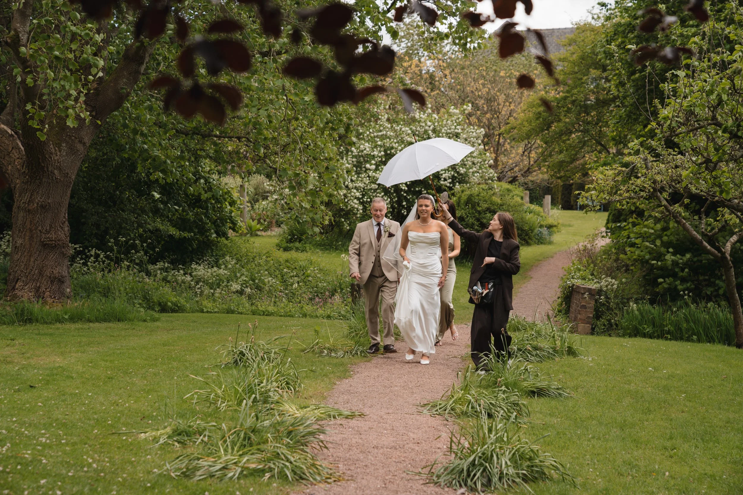 A wedding scene in a garden with a bride, groom, and two women walking on a garden path. One woman is holding a white umbrella over the bride. The garden is lush with green trees, bushes, and white flowering plants.