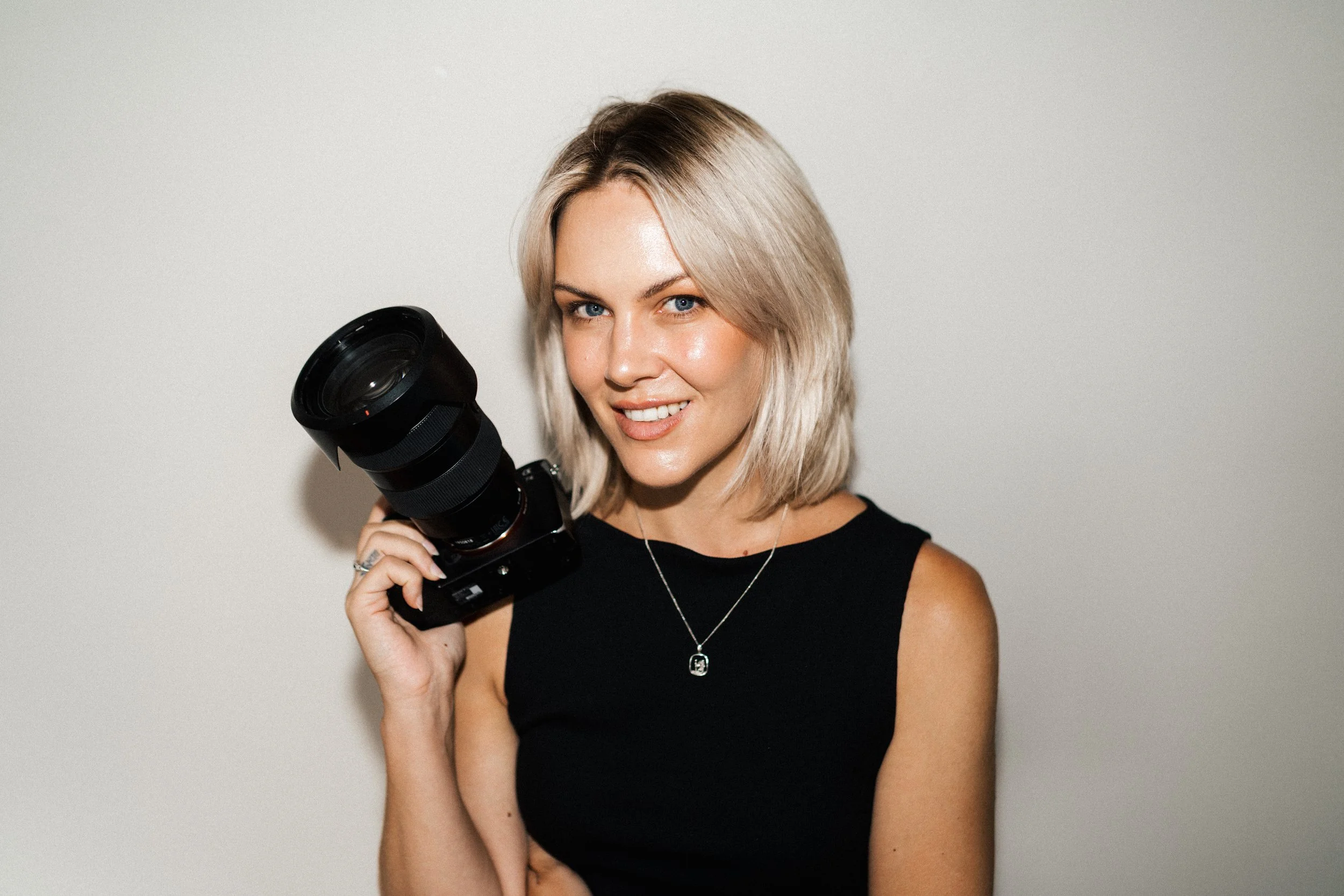 A smiling woman with blond hair holding a camera, wearing a black sleeveless top and a necklace, standing against a plain light background.