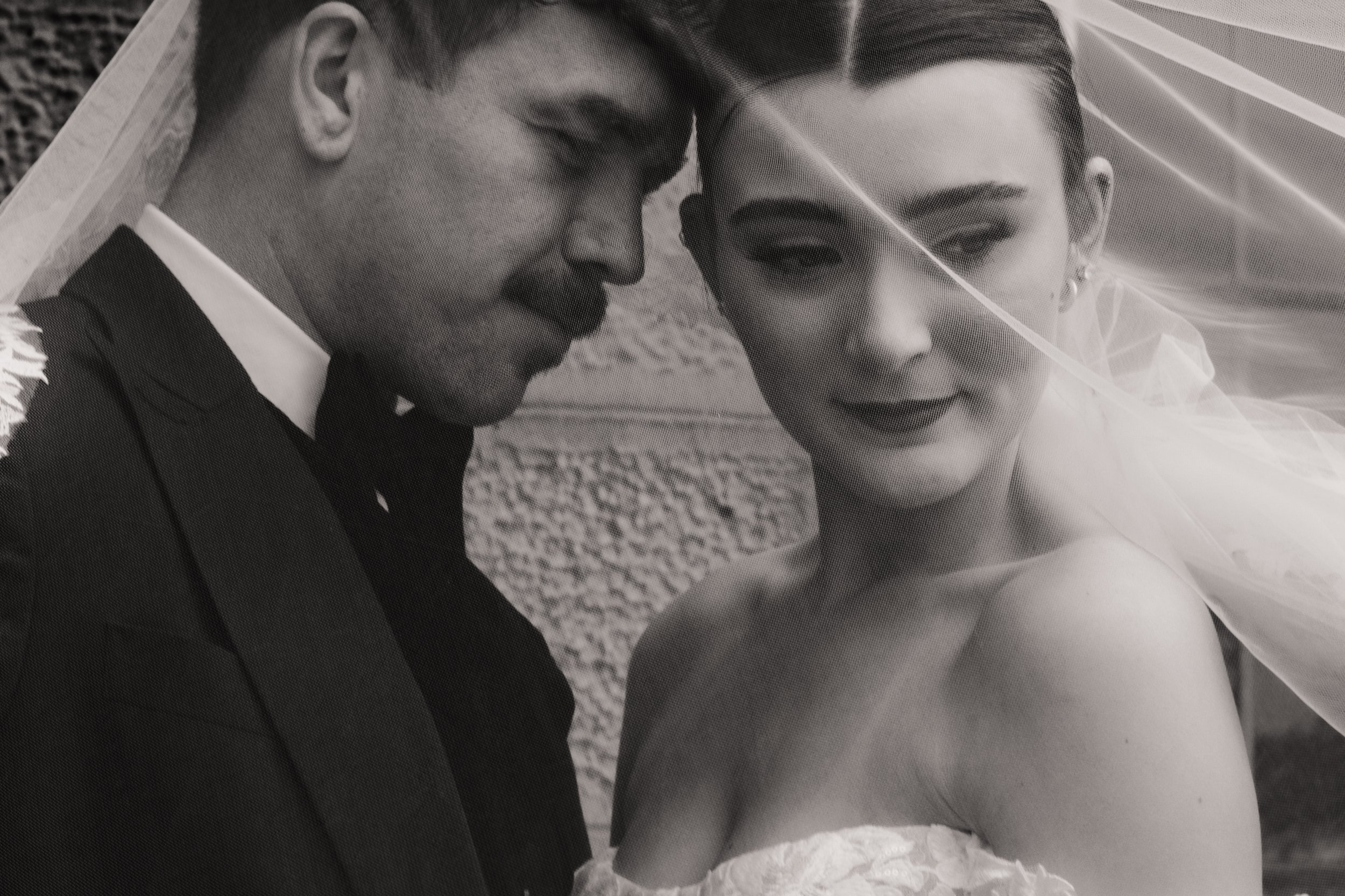 Black and white photo of a man and a woman close together, with the man leaning his forehead against the woman's forehead. The woman is wearing a bridal veil and earrings, and has makeup on. They both have their eyes closed, appearing to share an intimate moment.
