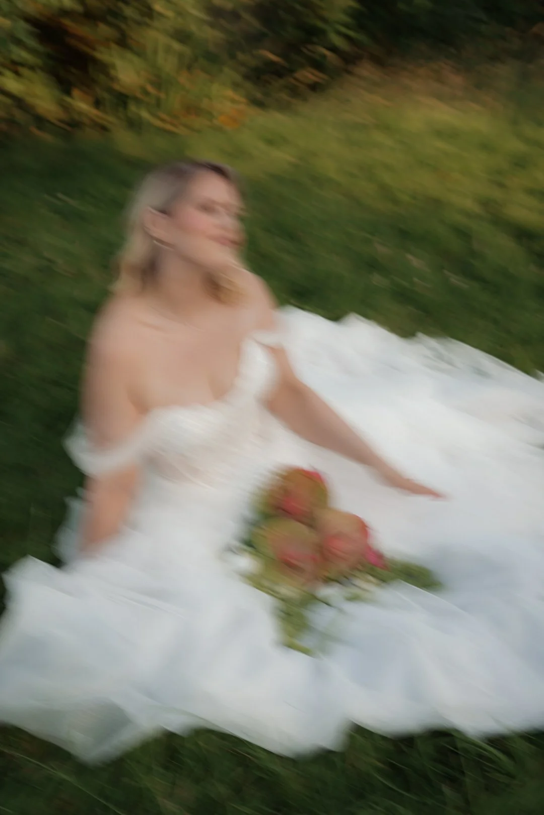 A woman in a white dress with off-the-shoulder sleeves sitting on a white blanket outdoors, with a bouquet of flowers in front of her, surrounded by greenery.