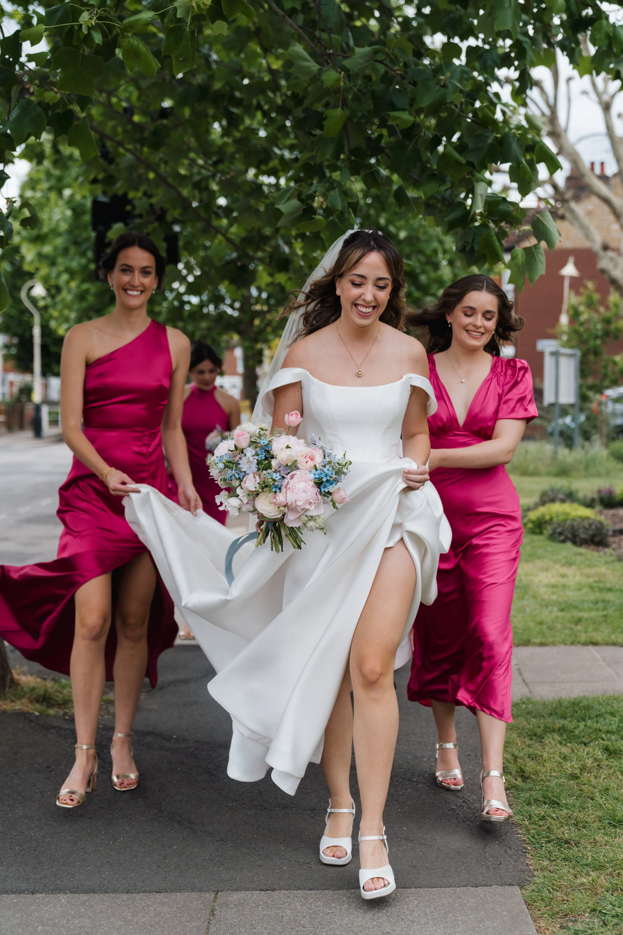 A bride in a white wedding dress walking outdoors on a sidewalk, smiling, accompanied by four bridesmaids in bright pink dresses. The bride is holding a bouquet of flowers and lifting her dress slightly. The scene is surrounded by green trees and bui