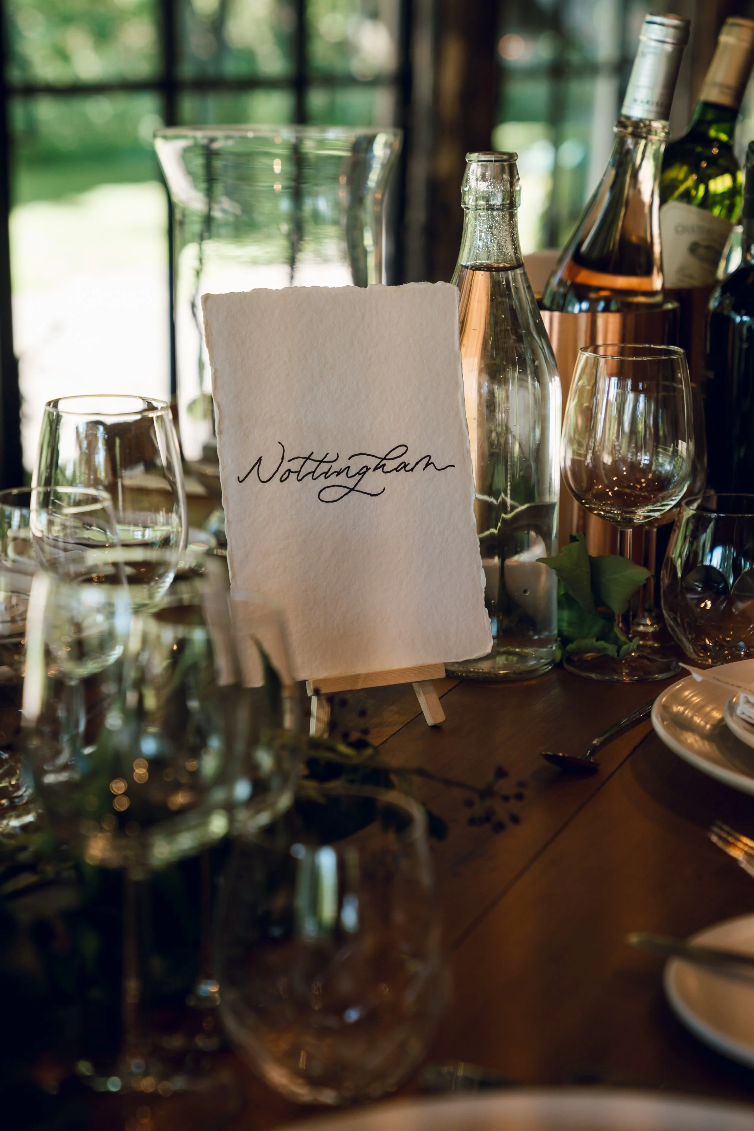 A table set with glassware, a water pitcher, bottles, a small easel with a card that has ‘Nottingham’ handwritten on it, and some greenery, inside a room with large windows showing greenery outside.