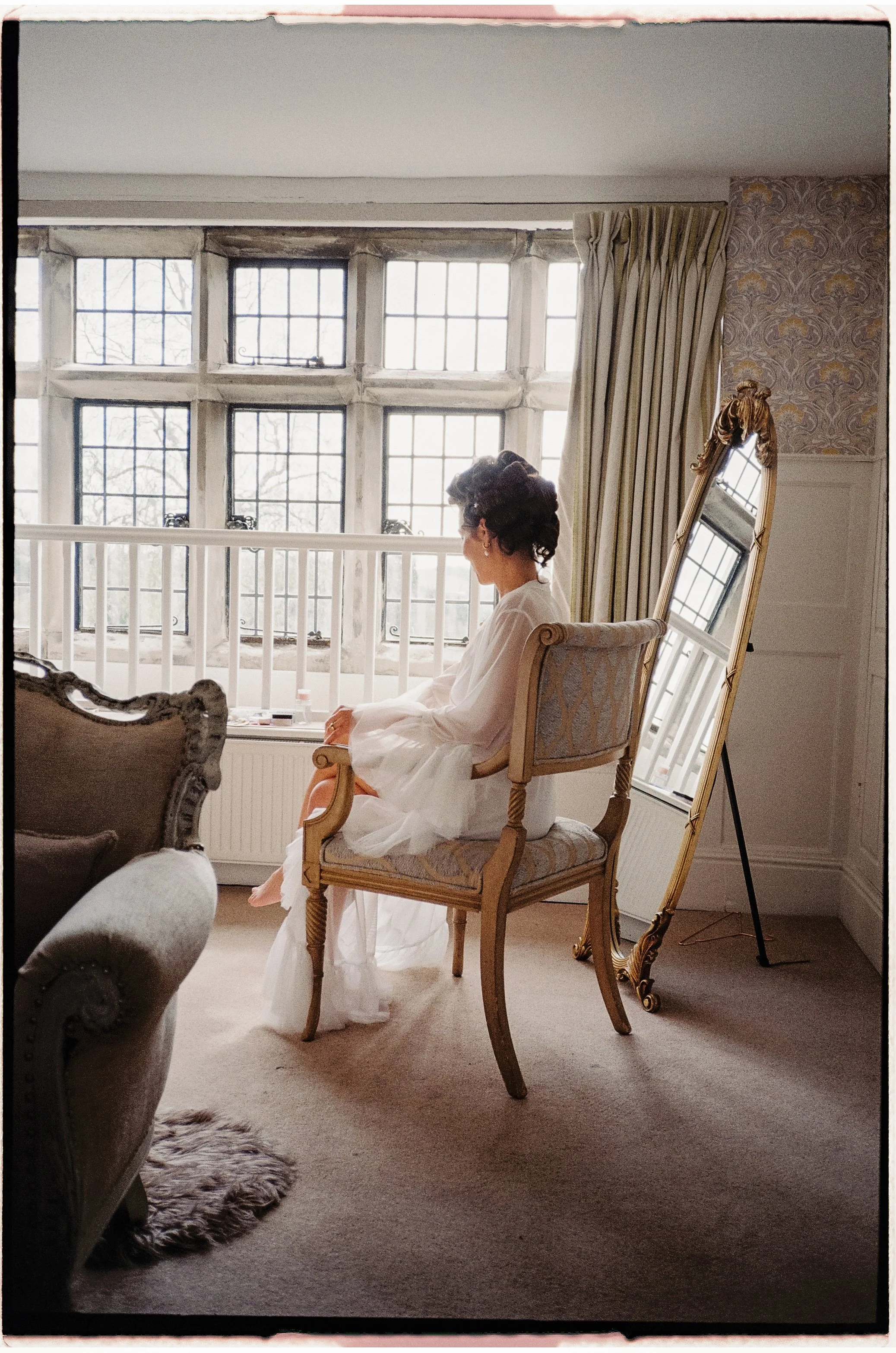 A woman with dark curly hair sitting on a vintage wooden chair in a room with large multi-pane windows and patterned wallpaper.