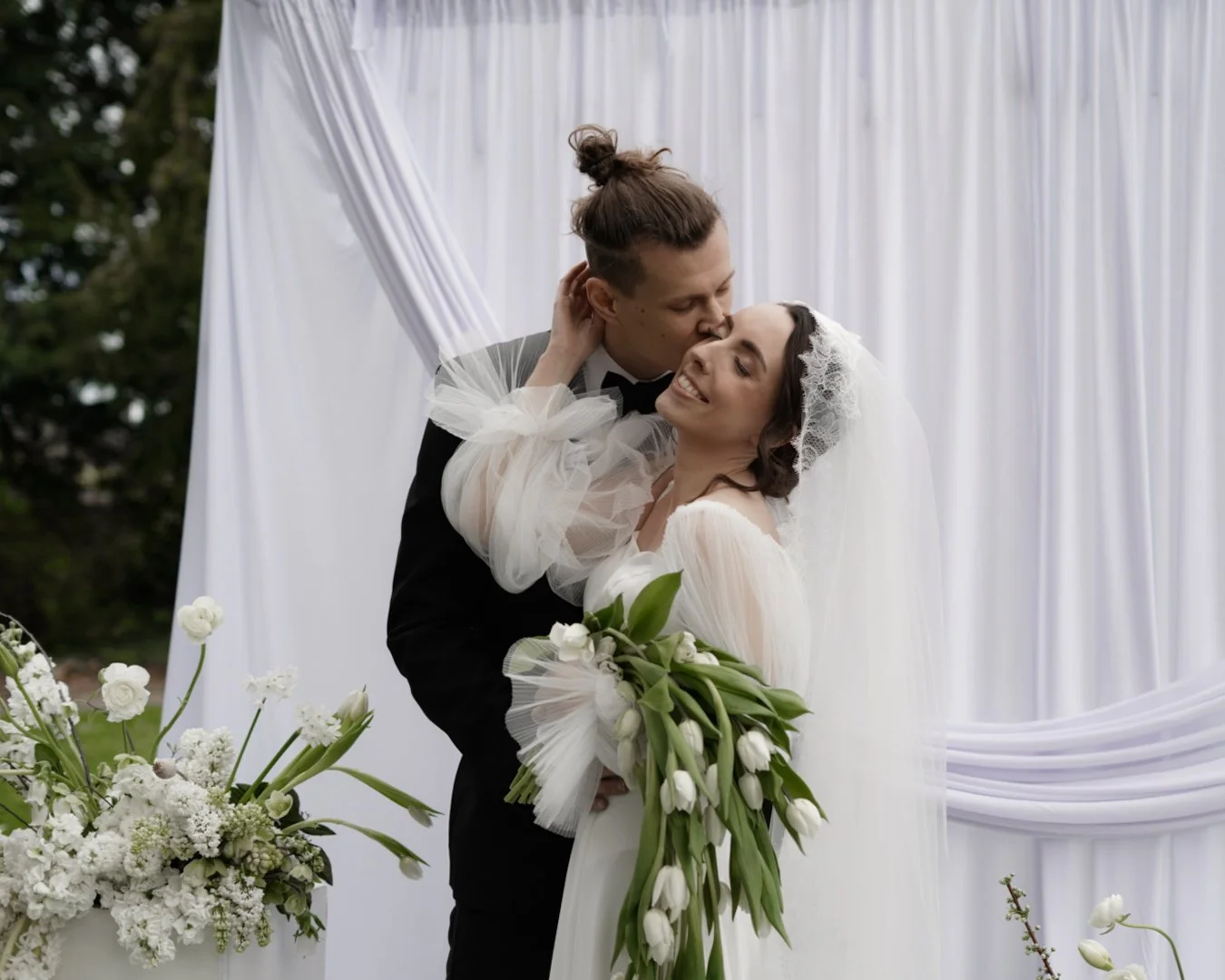 A newlywed couple sharing a romantic kiss outdoors, with the groom in a tuxedo and the bride in a white wedding dress holding a bouquet of white tulips and greenery, standing in front of white drapery and floral arrangements.