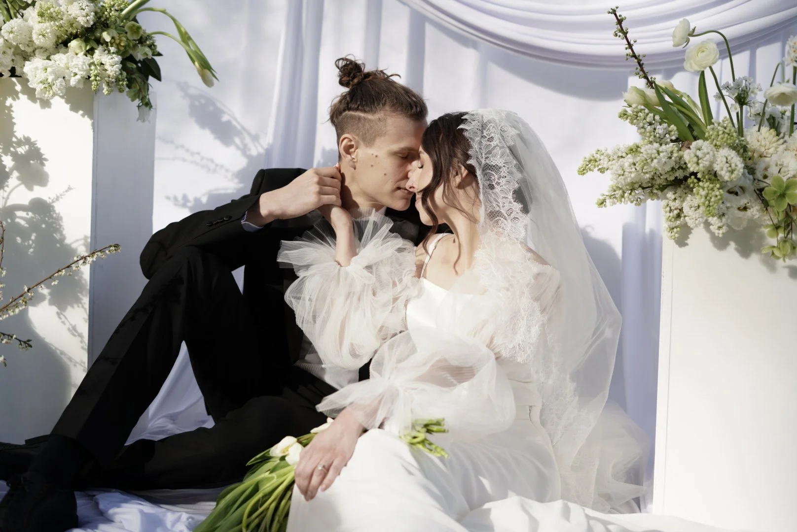 A bride and groom share a romantic kiss during their wedding ceremony, surrounded by white flowers and draped white fabric.