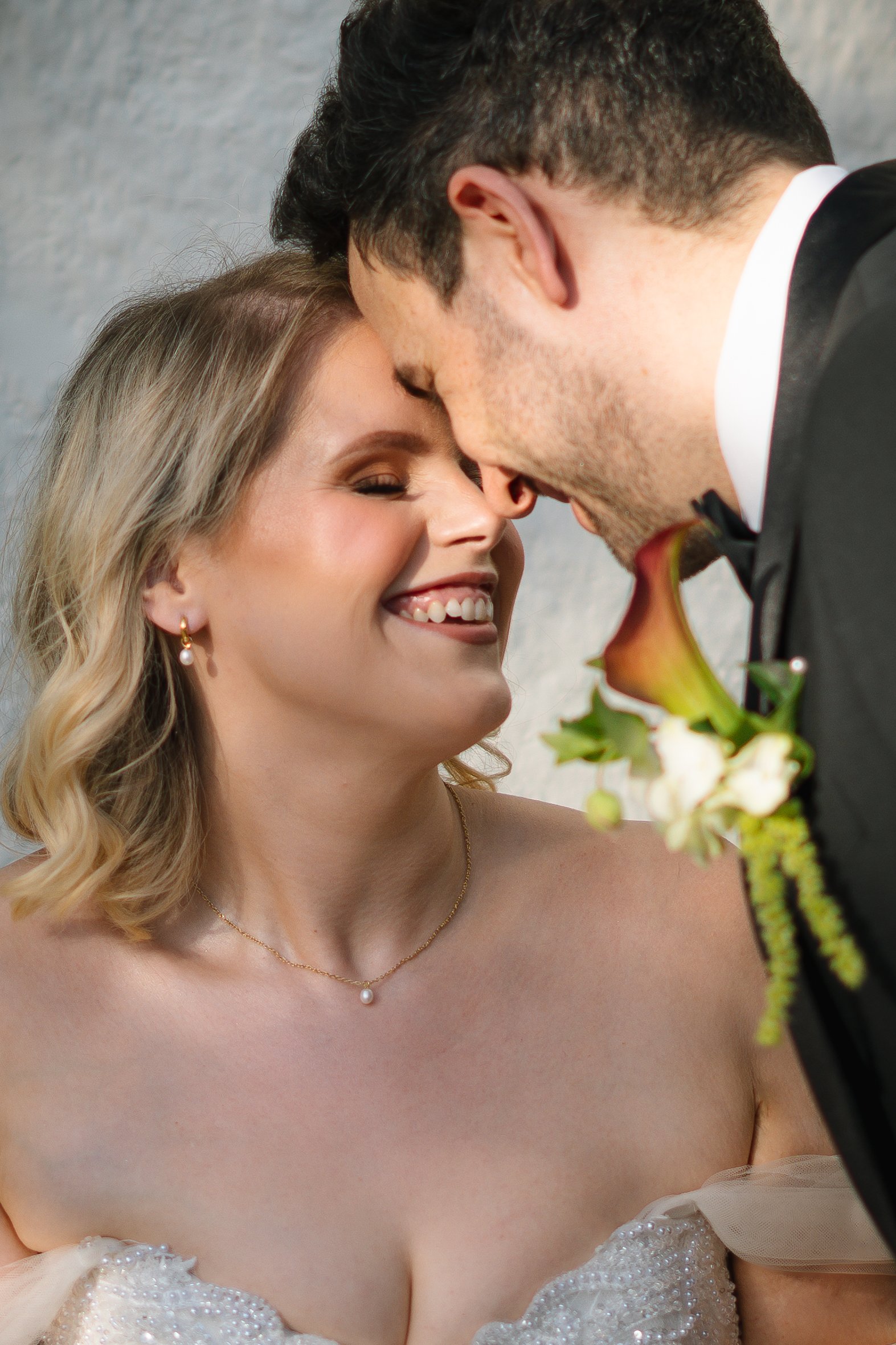 A man and woman are touching foreheads in a close, intimate moment. The woman is smiling with her eyes closed, wearing a pearl earring, a delicate necklace, and a white dress with pearl embellishments. The man is wearing a black tuxedo with a bowtie,
