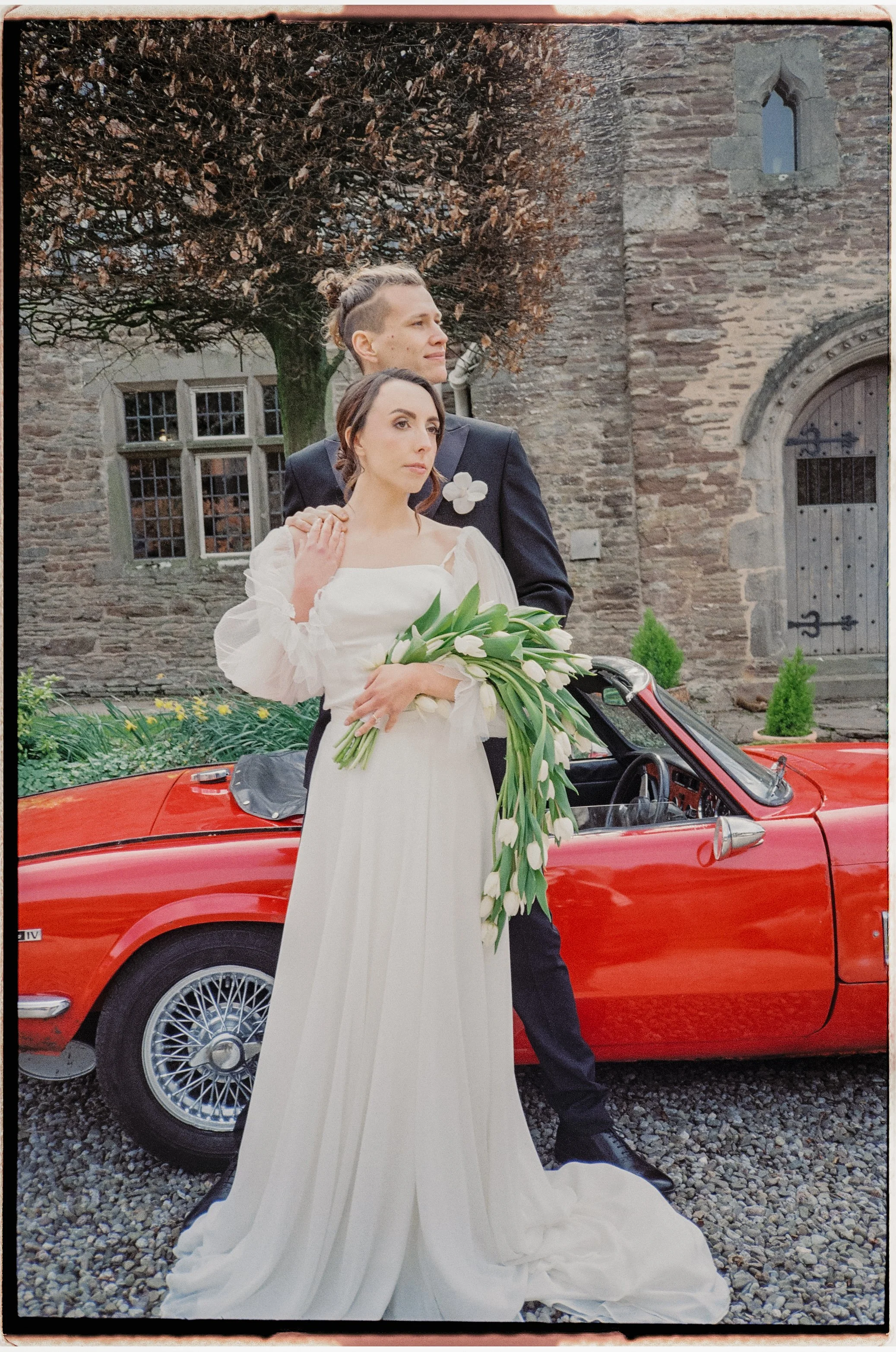 A couple in wedding attire standing next to a red vintage car in front of a stone building with trees.