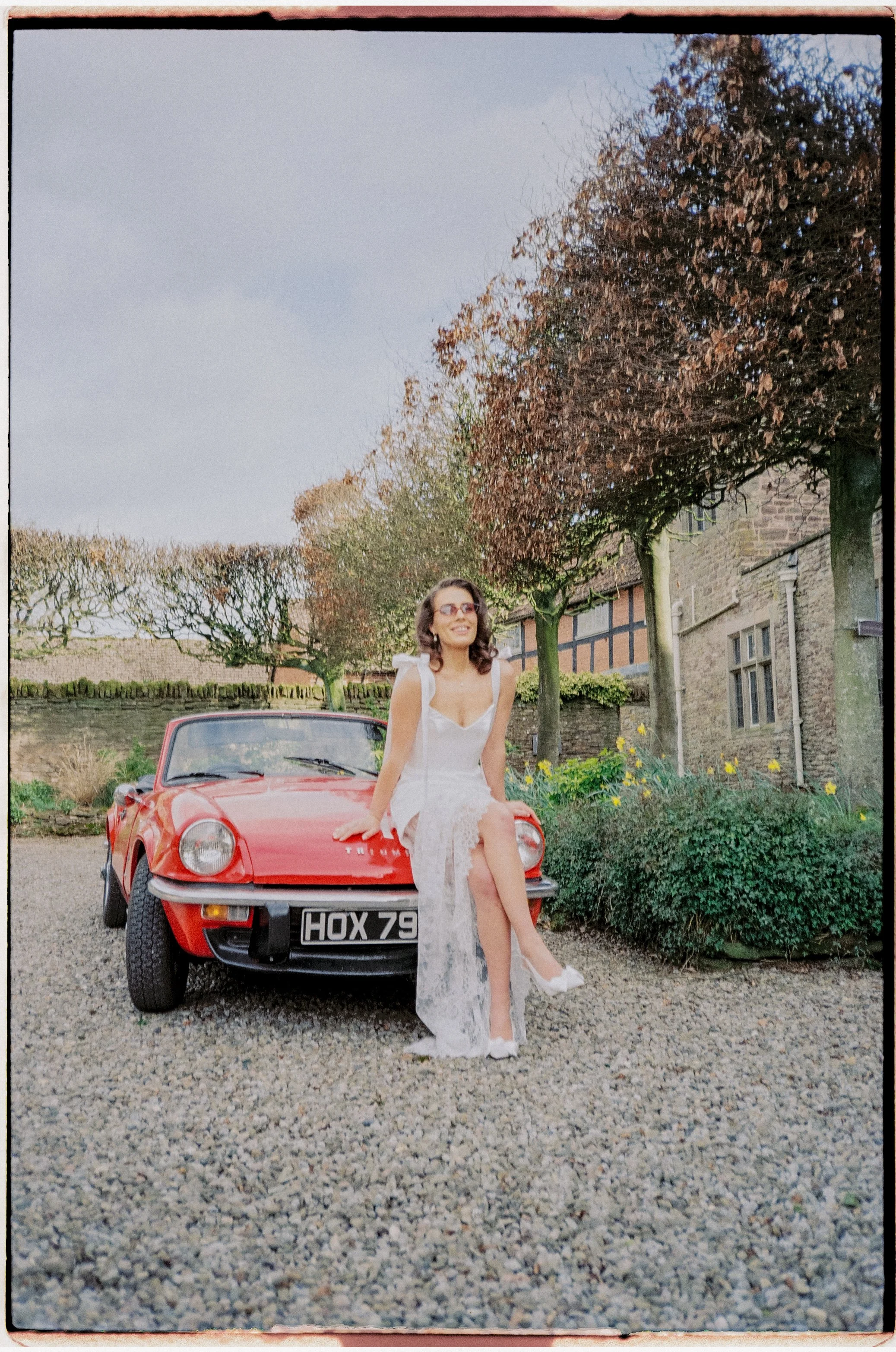 A woman in a white dress sitting on the hood of a red vintage car in a garden setting with trees and a stone building in the background.