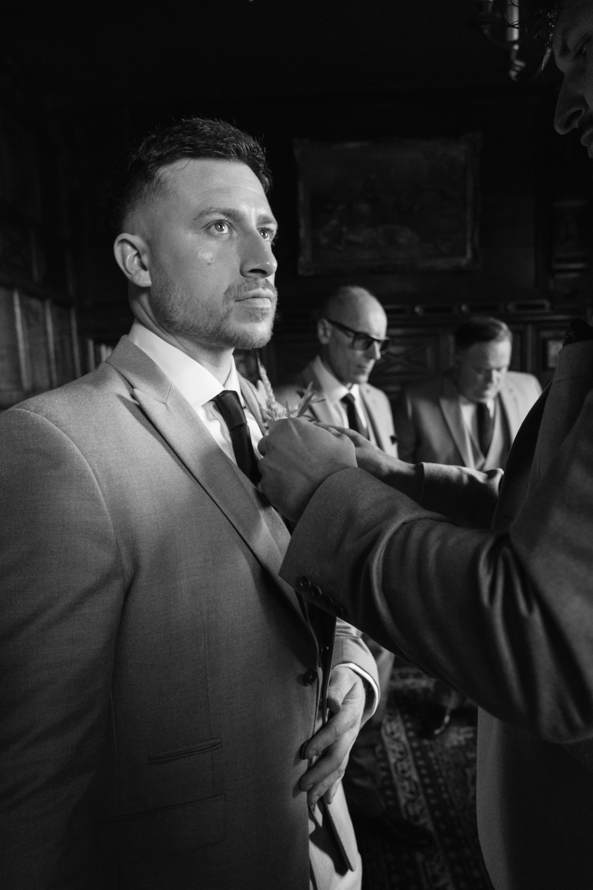 A group of men in formal suits standing indoors, with one man prominently in the foreground gazing forward, and others behind him, some with glasses, engaged in a formal setting.