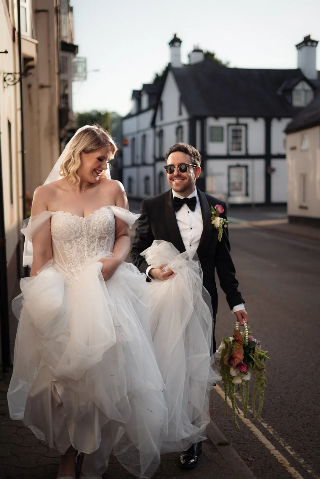 A newlywed couple walking on a street in wedding attire, the bride in a white gown and veil, and the groom in a black tuxedo with sunglasses, holding a bouquet, and smiling.