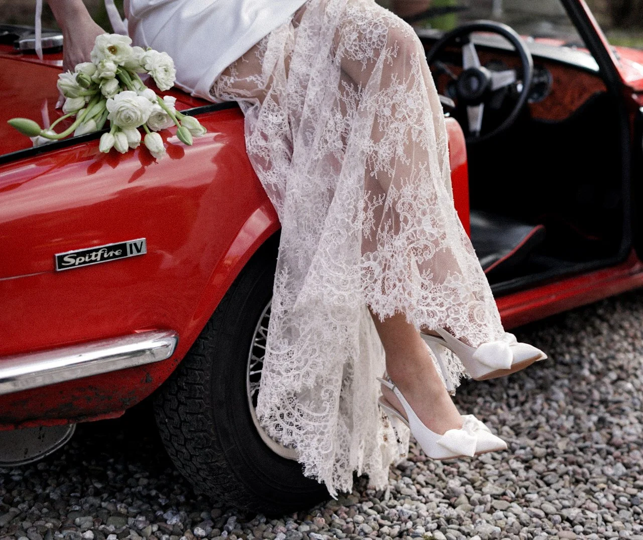 Close-up of a bride's legs and shoes hanging out of a red vintage Spitfire IV car, wearing a lace wedding dress and white high heels with bows, with a bouquet resting on the car's hood.