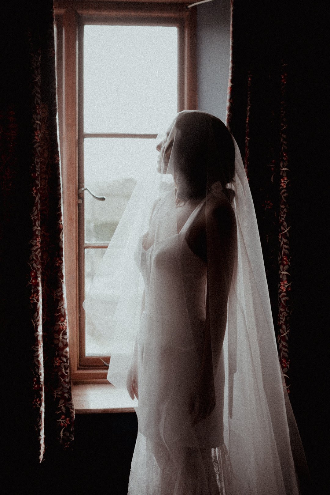 Woman in wedding dress and veil standing by window with curtains on each side, backlit by natural light.
