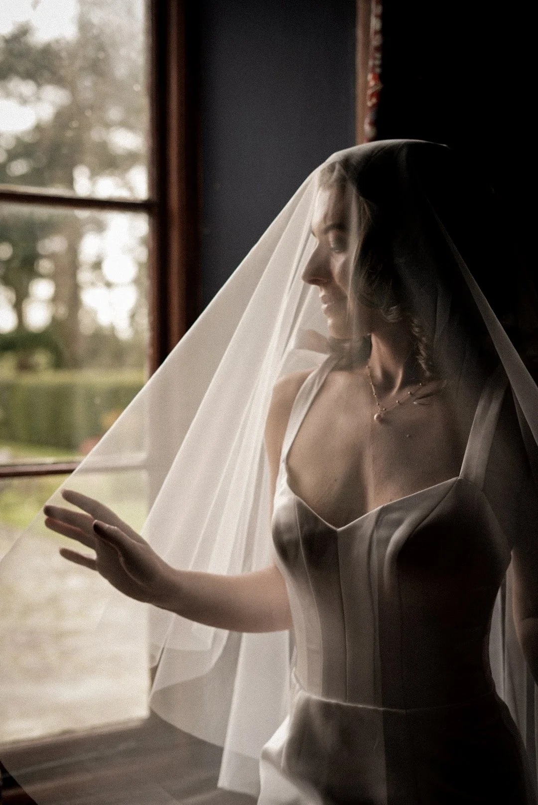 A bride looking out a window with sheer veil over her face, wearing a satin wedding dress with thin straps, inside a dimly lit room.
