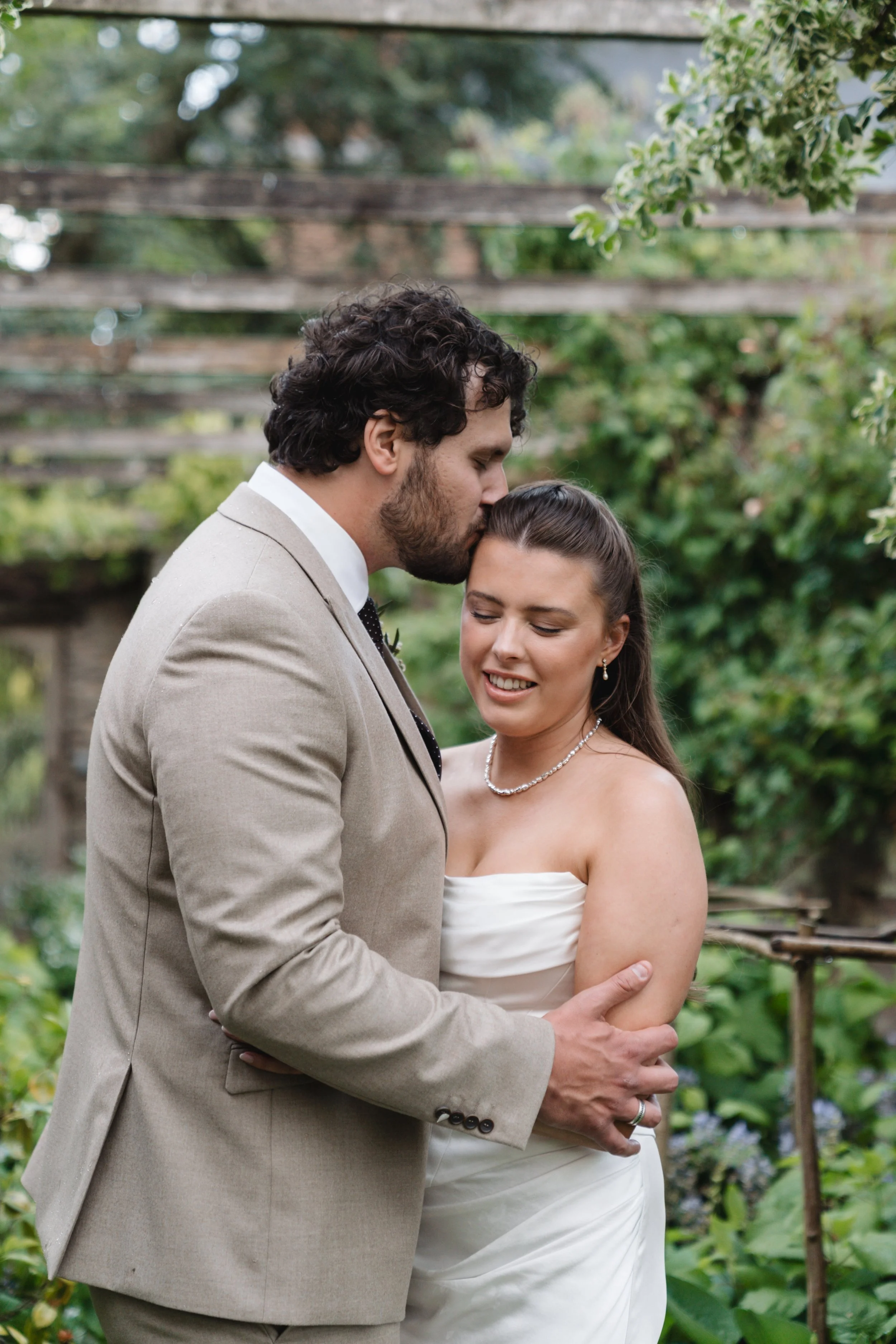 A couple in wedding attire sharing an intimate moment outdoors, with the man kissing the woman on her forehead, surrounded by lush greenery and wooden beams in the background.