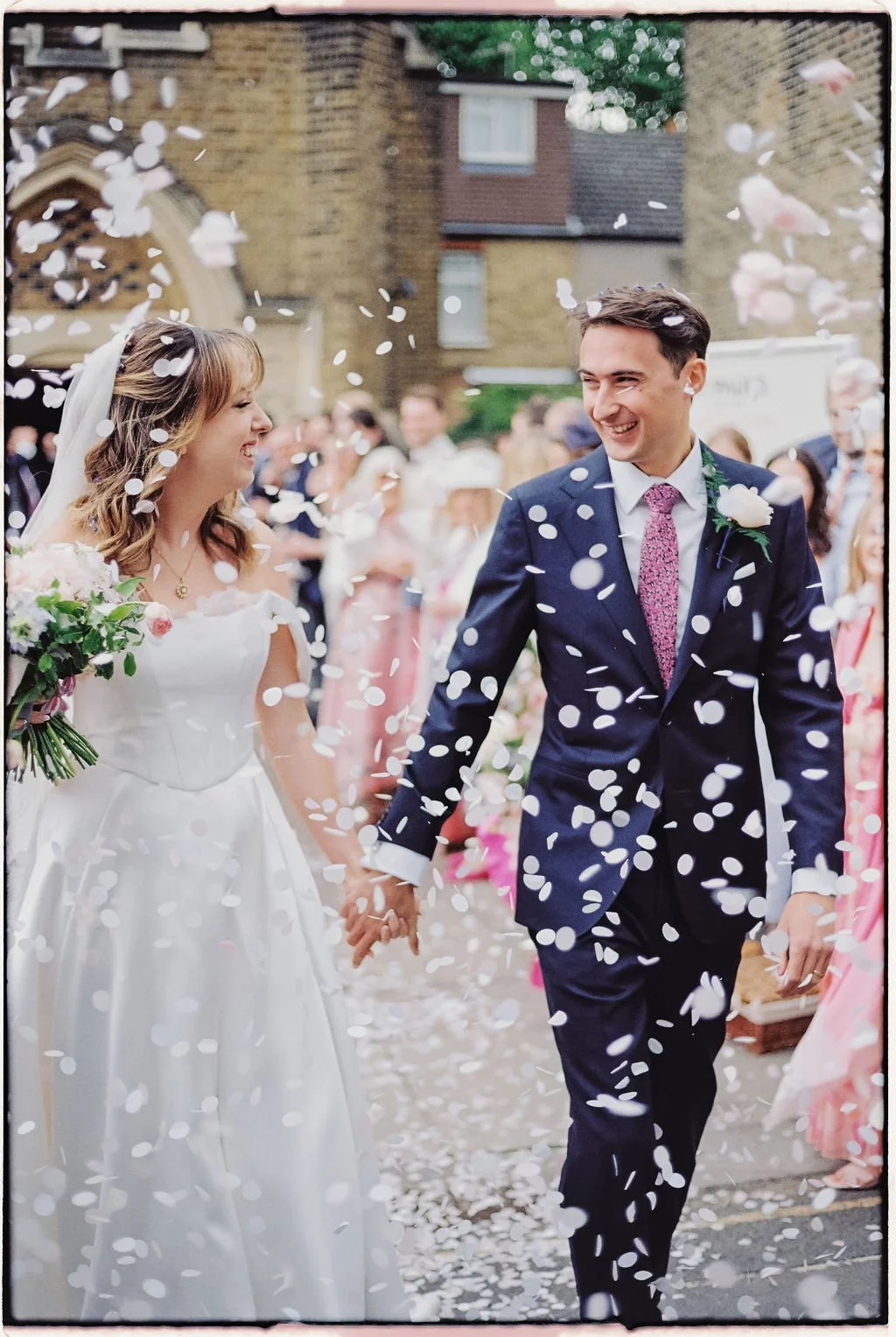 A bride and groom walking hand-in-hand outside church, surrounded by guests and falling confetti, celebrating their wedding. Capture on 35mm film. 35mm film photography.