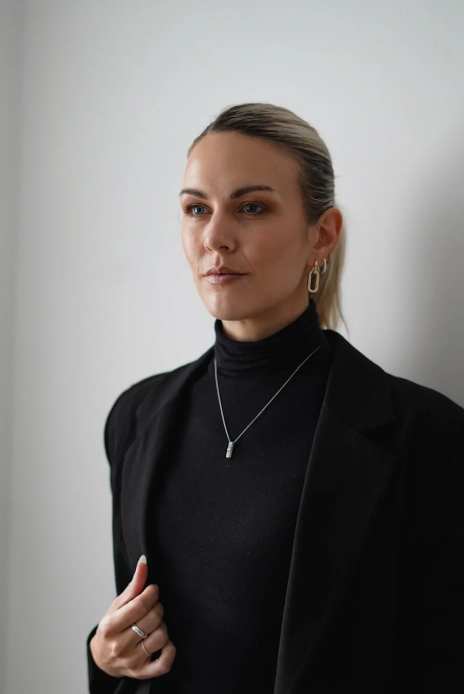 A woman with blonde hair tied back, wearing a black turtleneck, black blazer, and jewelry including earrings, a necklace, and a ring, standing against a plain light-colored wall.