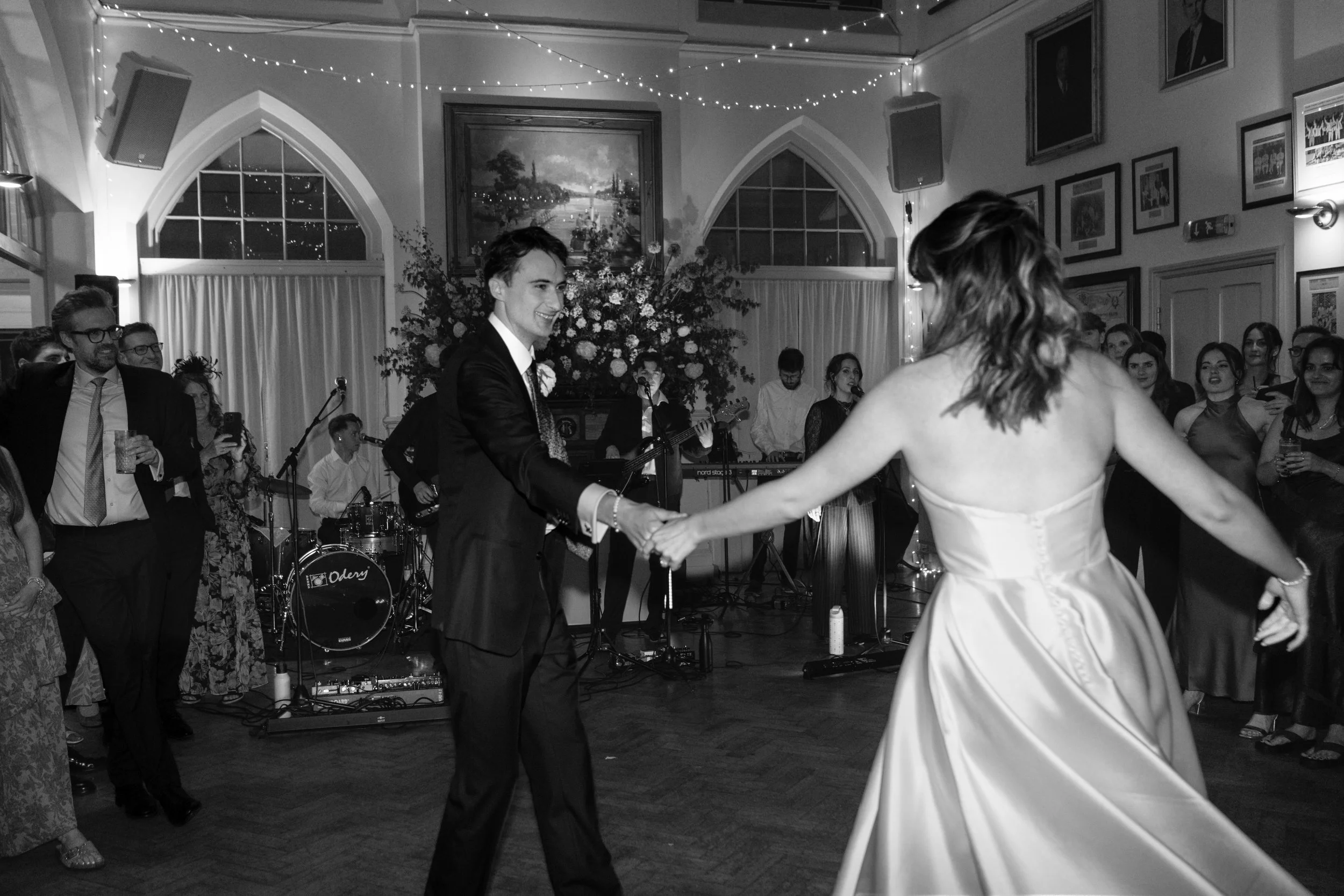 A bride and groom dancing at their wedding reception, surrounded by guests in a decorated hall with a band playing music.