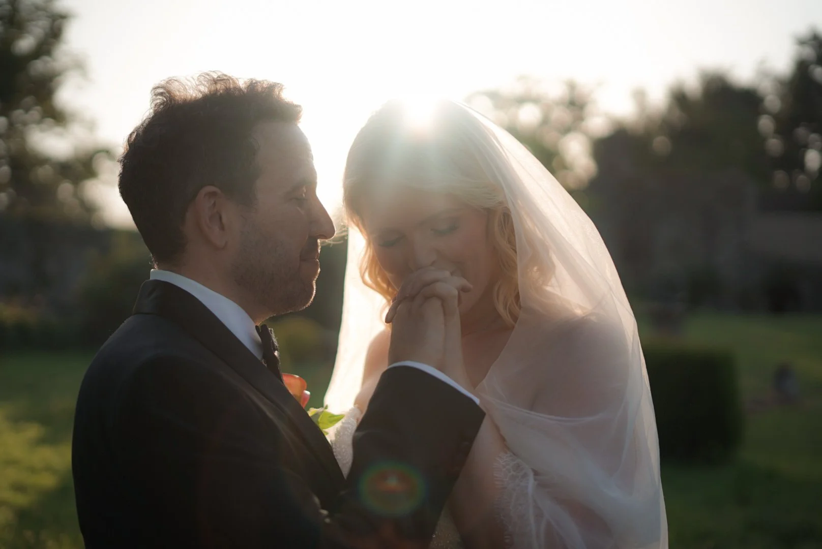 A groom and bride holding hands and touching foreheads outdoors during sunset, with the sun shining behind them.